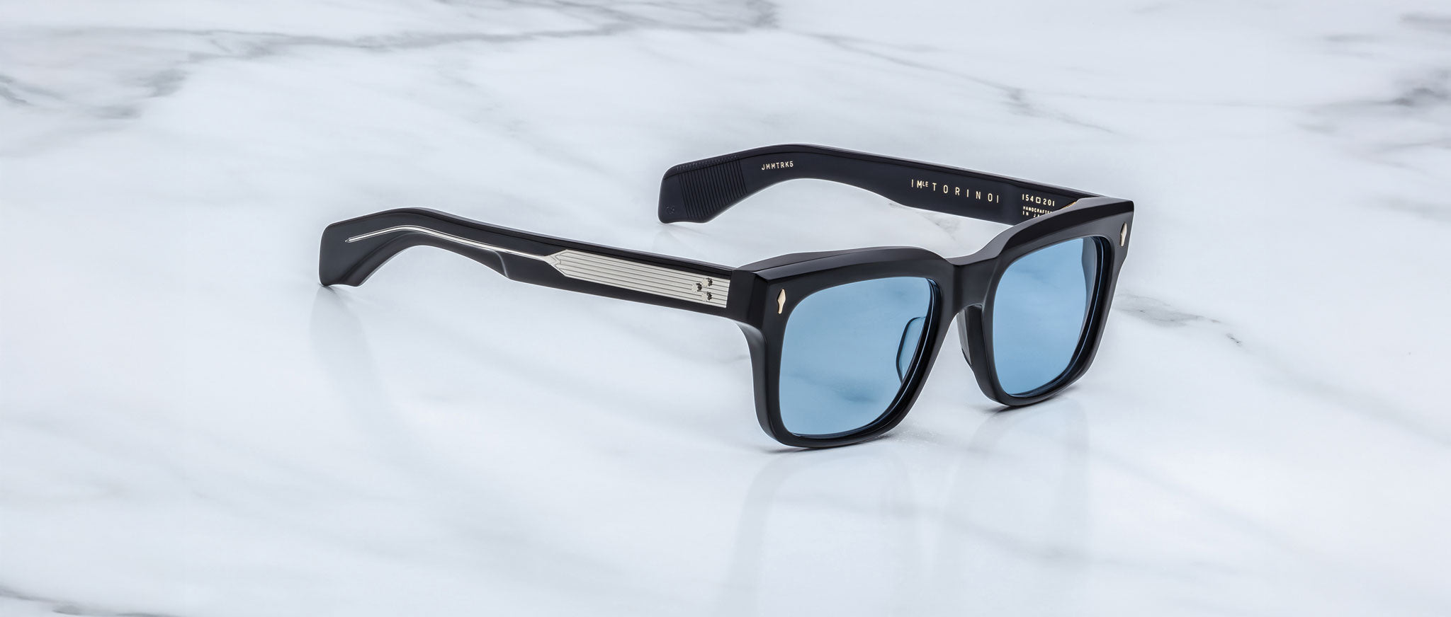 The Jacques Marie Mage Torino sunglasses by Jacques Marie Mage feature bold black rectangular acetate frames, blue-tinted lenses, and silver arm detailing, elegantly displayed on a white marble surface.