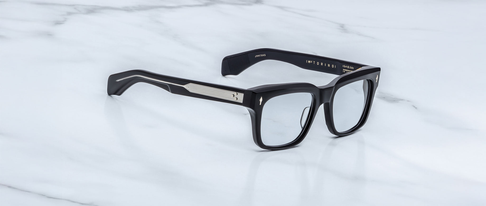 Jacques Marie Mage Torino eyeglasses by Jacques Marie Mage feature bold black rectangular acetate frames with silver arm detailing, elegantly displayed on a white marble surface.