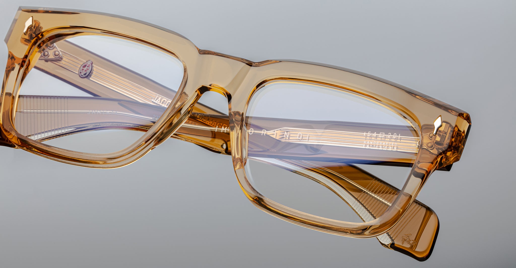 The Jacques Marie Mage Torino, bold rectangular eyeglass frames in amber transparent acetate by Jacques Marie Mage, rest on a light gray surface—a striking example of handcrafted acetate eyewear.