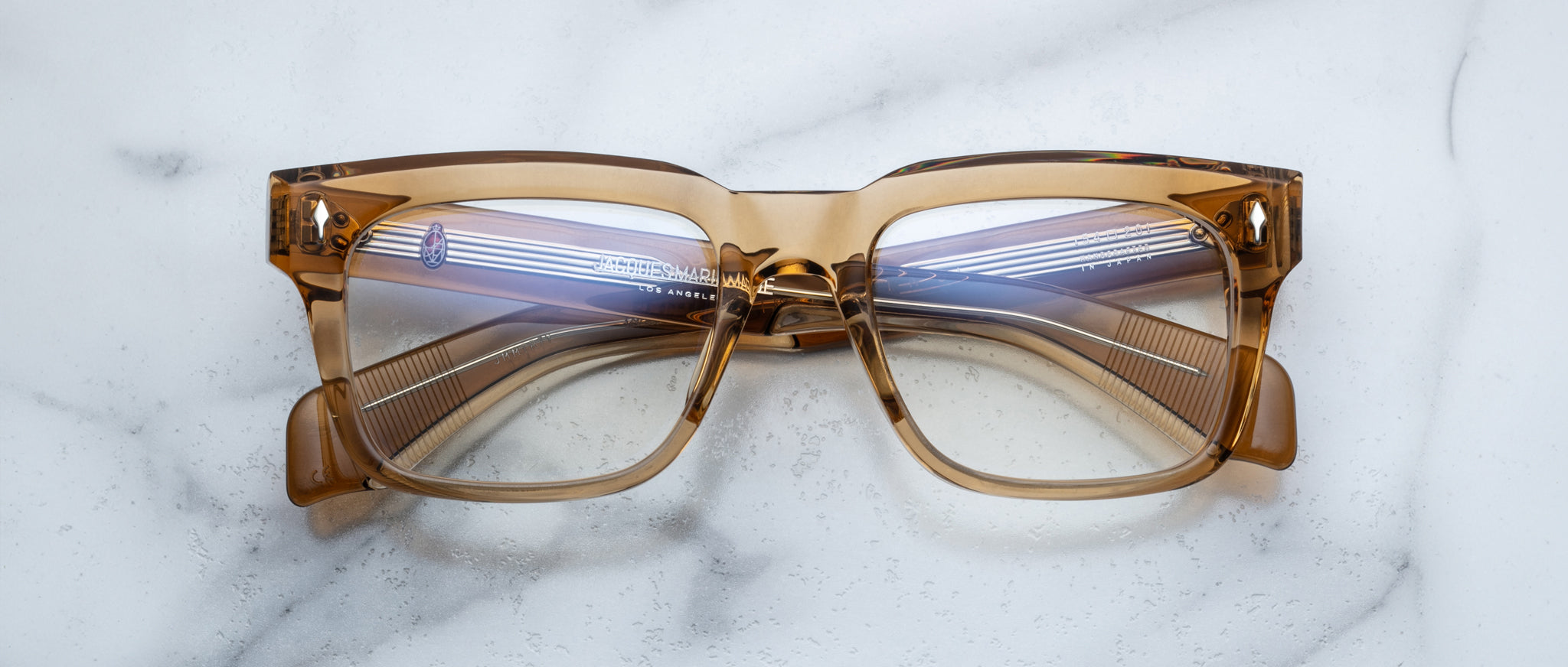 The Jacques Marie Mage Torino eyeglasses feature bold, handcrafted acetate rectangular frames with clear lenses, displayed on a white marble surface.
