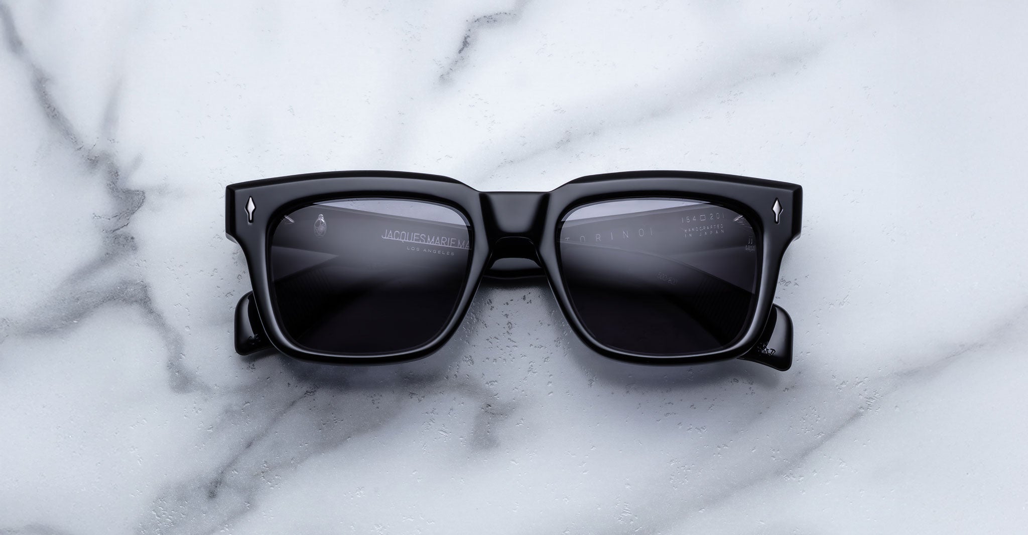 Jacques Marie Mage Torino sunglasses by Jacques Marie Mage, featuring bold black rectangular acetate frames with dark lenses, are displayed on a white marble surface.