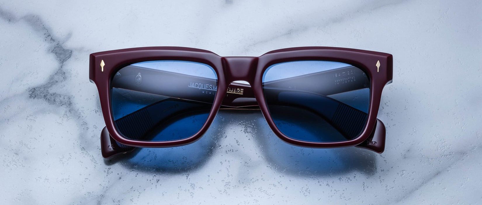 The Jacques Marie Mage Torino sunglasses by Jacques Marie Mage feature bold square frames in dark red acetate with blue lenses, elegantly displayed on a white marble surface.