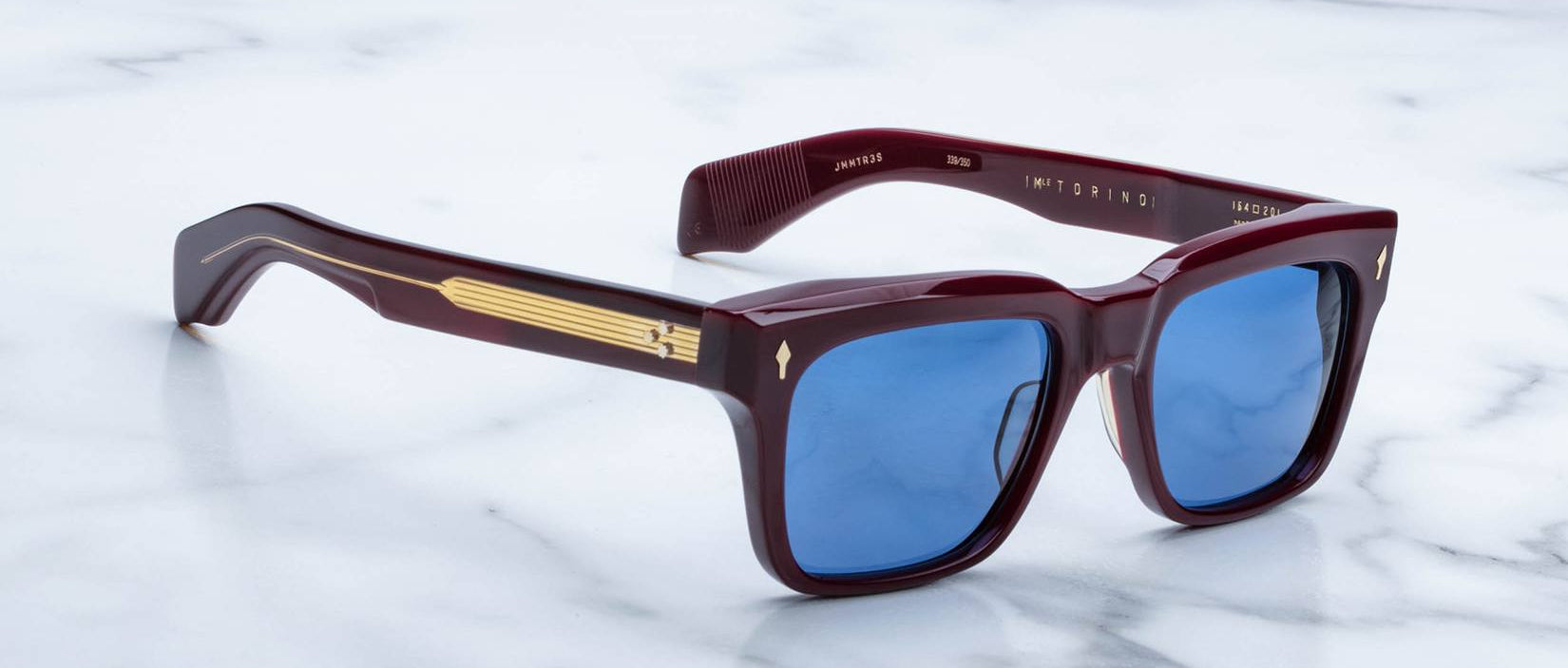 A pair of Jacques Marie Mage Torino square burgundy sunglasses with blue lenses and gold temple detailing—bold frames by Jacques Marie Mage—rest on a white marble surface.