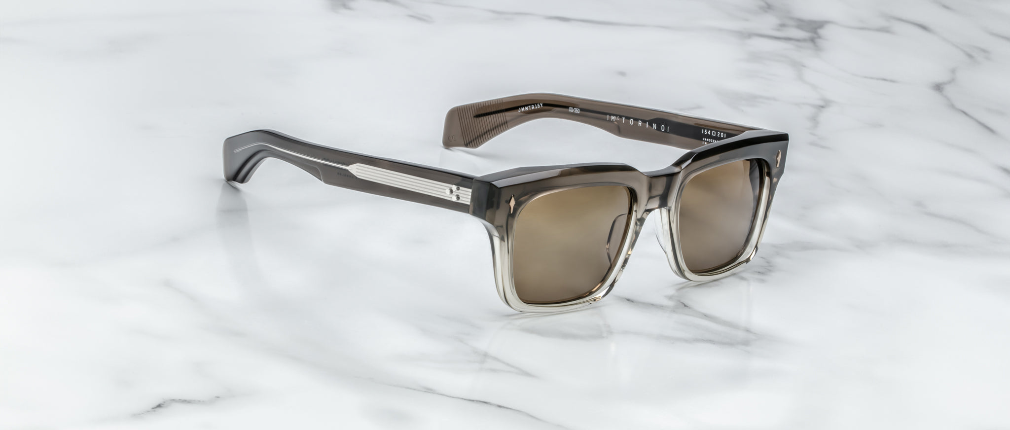 A pair of Jacques Marie Mage Torino sunglasses by Jacques Marie Mage, featuring bold rectangular frames and tinted lenses, rests on a white marble surface.