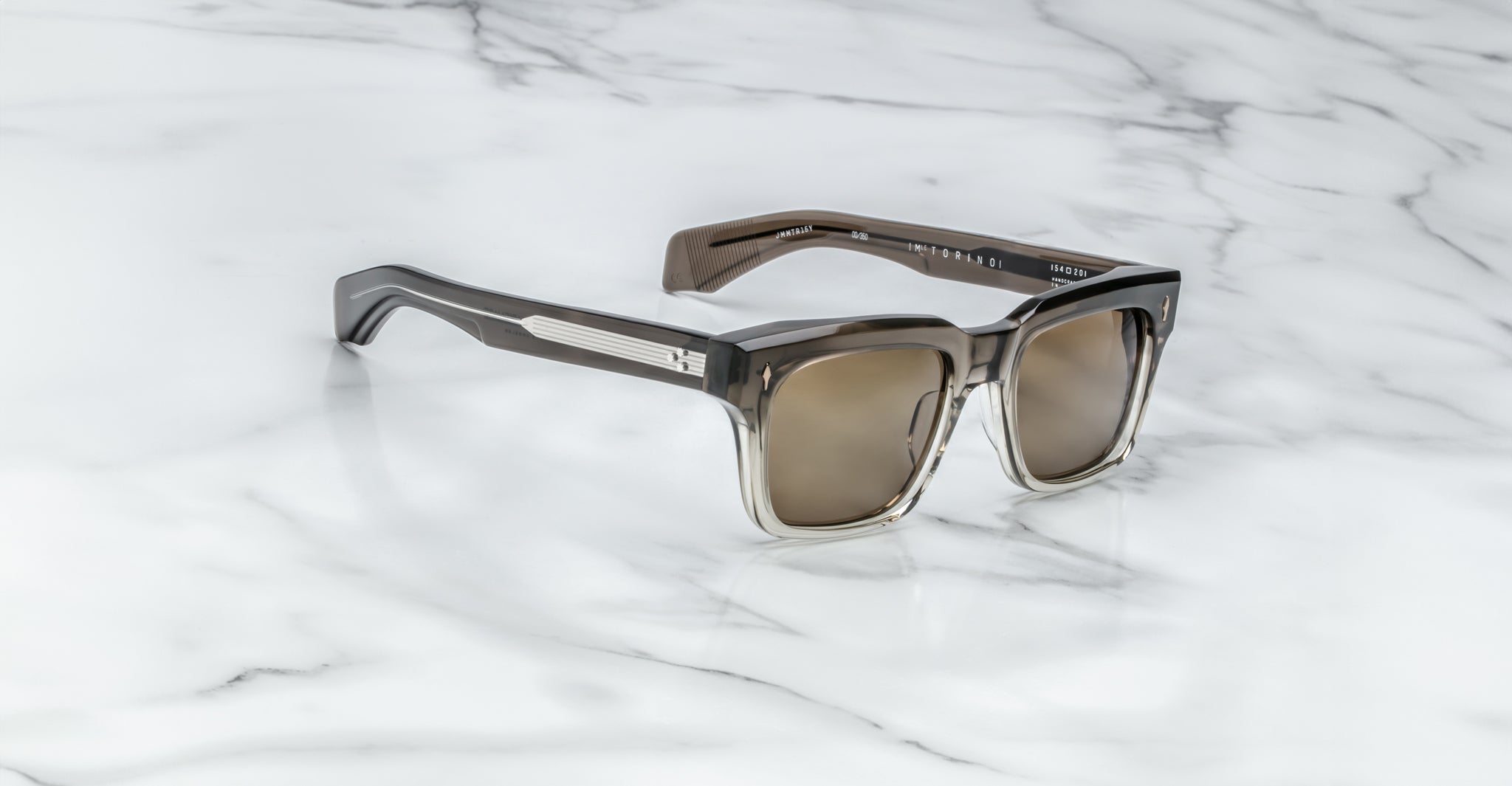 A pair of Jacques Marie Mage Torino sunglasses by Jacques Marie Mage, featuring bold rectangular frames and tinted lenses, rests on a white marble surface.