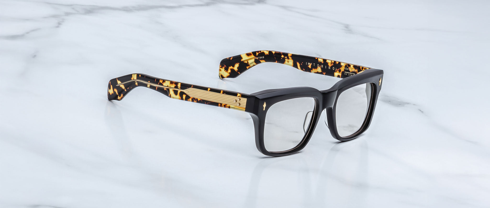 The Jacques Marie Mage Torino eyeglass frames feature bold rectangular black fronts and tortoiseshell-patterned arms, displayed on a white marble surface.