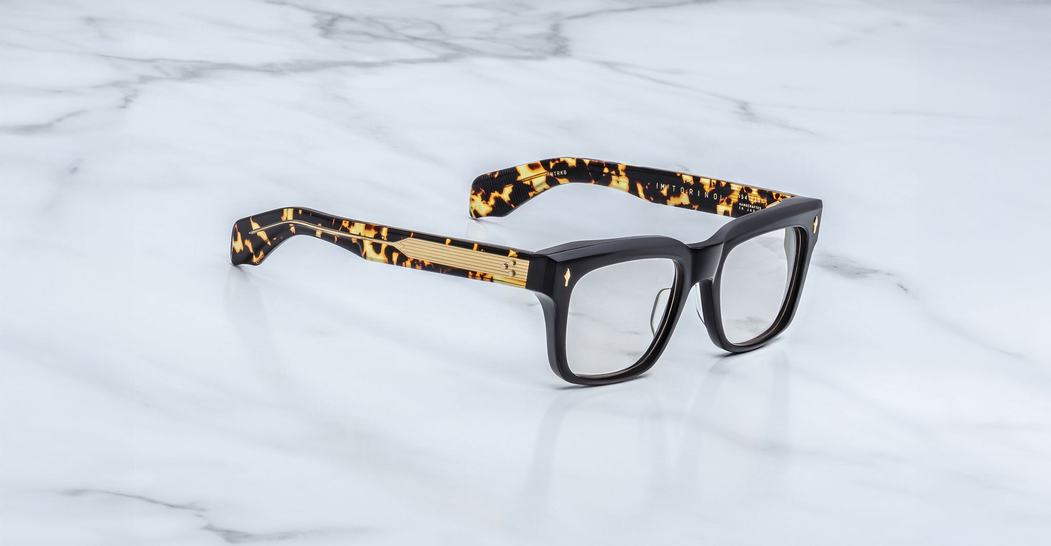 The Jacques Marie Mage Torino eyeglass frames feature bold rectangular black fronts and tortoiseshell-patterned arms, displayed on a white marble surface.