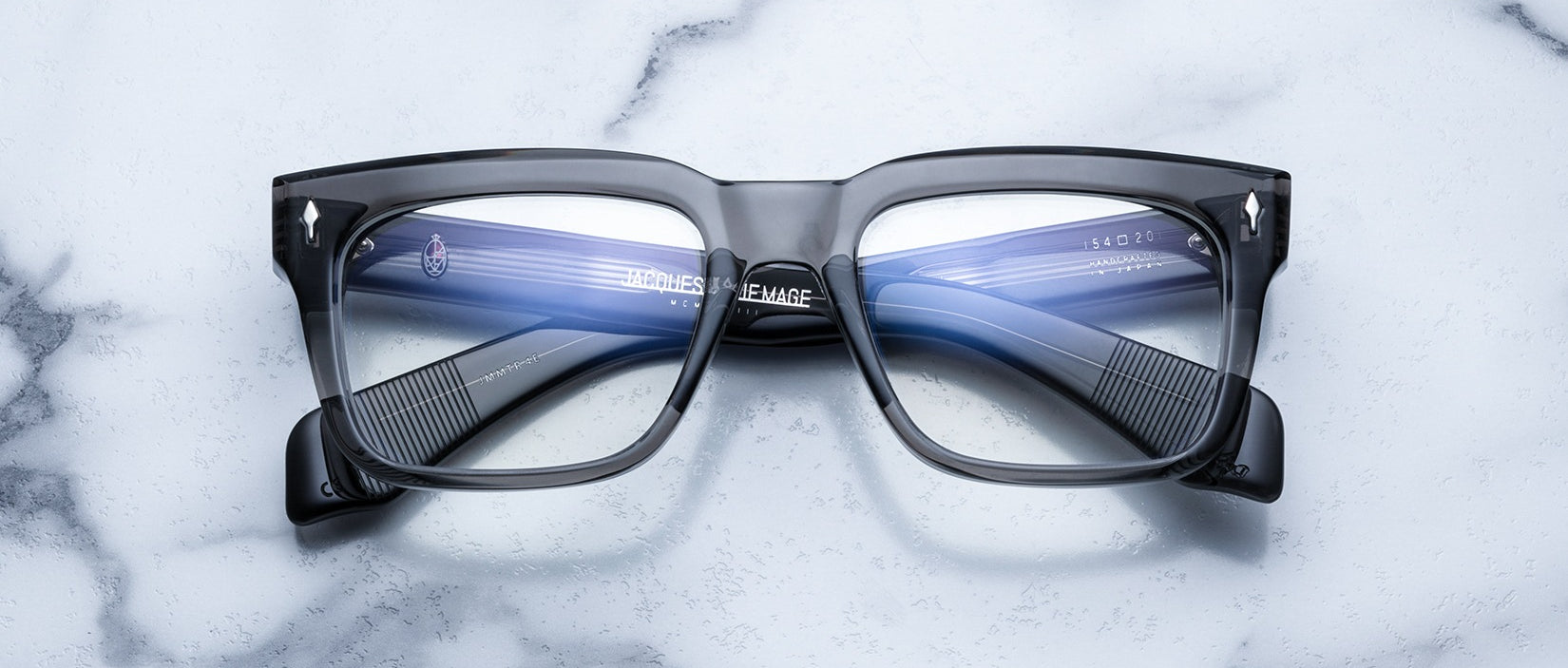 A pair of Jacques Marie Mage Torino rectangular eyeglass frames by Jacques Marie Mage, featuring blue-tinted lenses and handcrafted from premium acetate, displayed on a white marble surface.