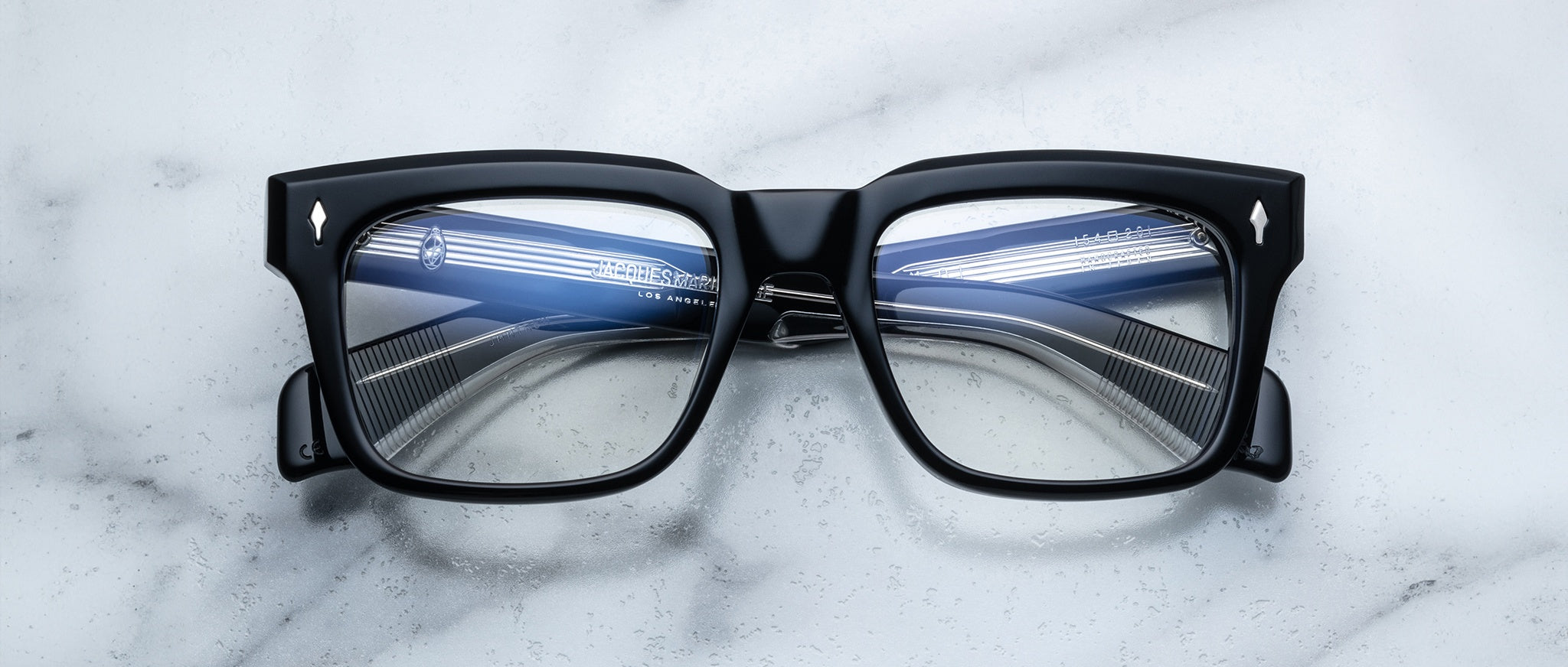 The Jacques Marie Mage Torino eyeglass frames by Jacques Marie Mage feature bold rectangular design with striped blue and white reflections on the lenses, displayed on a white marble surface.
