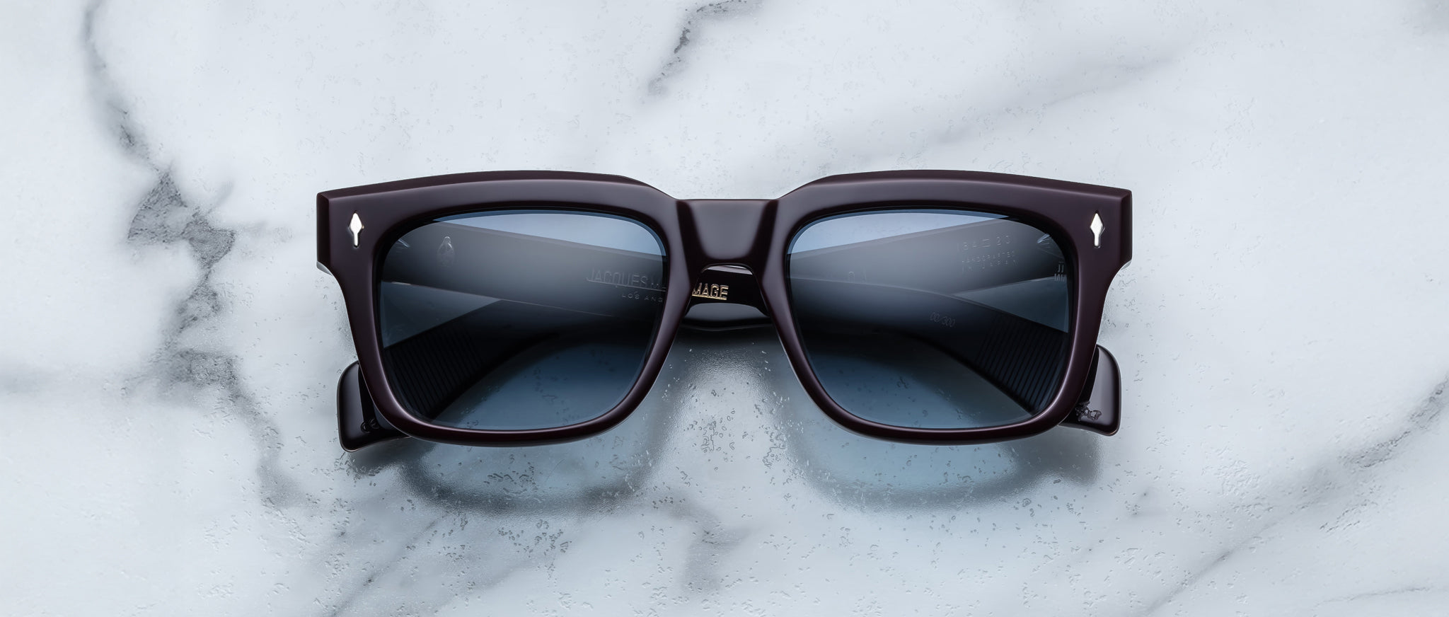 The Jacques Marie Mage Torino sunglasses by Jacques Marie Mage feature bold black rectangular frames and dark lenses, elegantly displayed on a white marble surface.