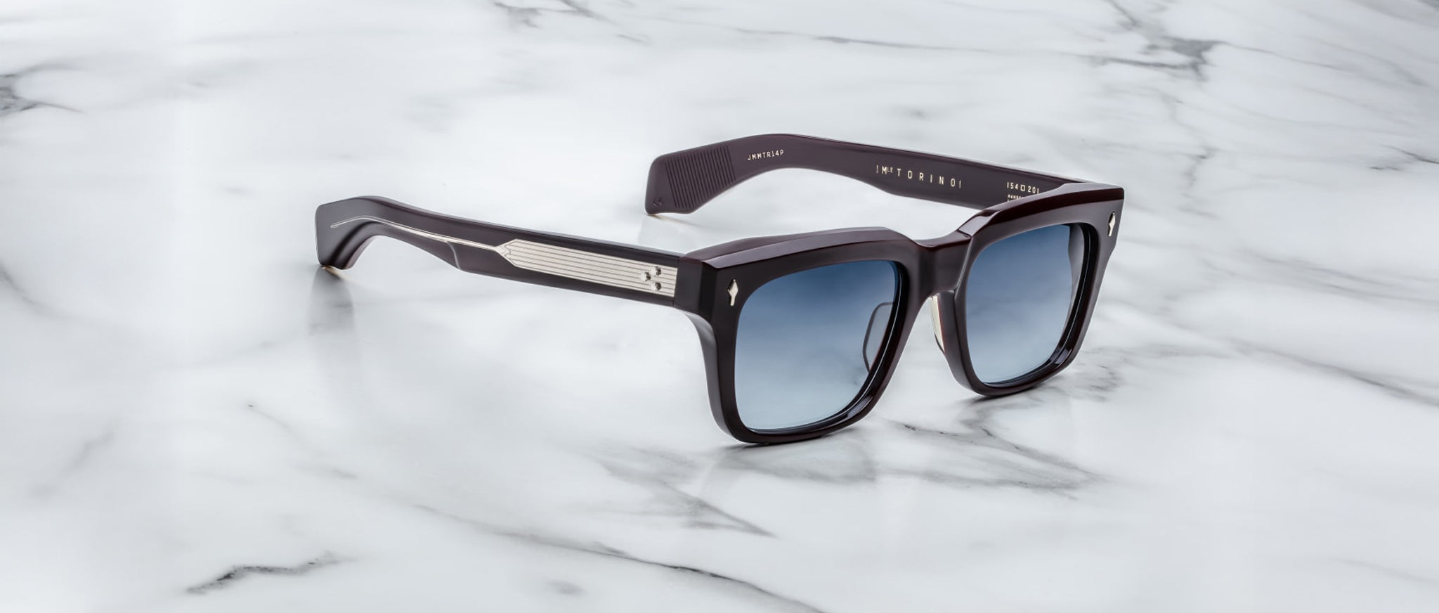 The Jacques Marie Mage Torino by Jacques Marie Mage features black handcrafted acetate rectangular frames, blue gradient lenses, and metallic arm accents, shown on a white marble surface.