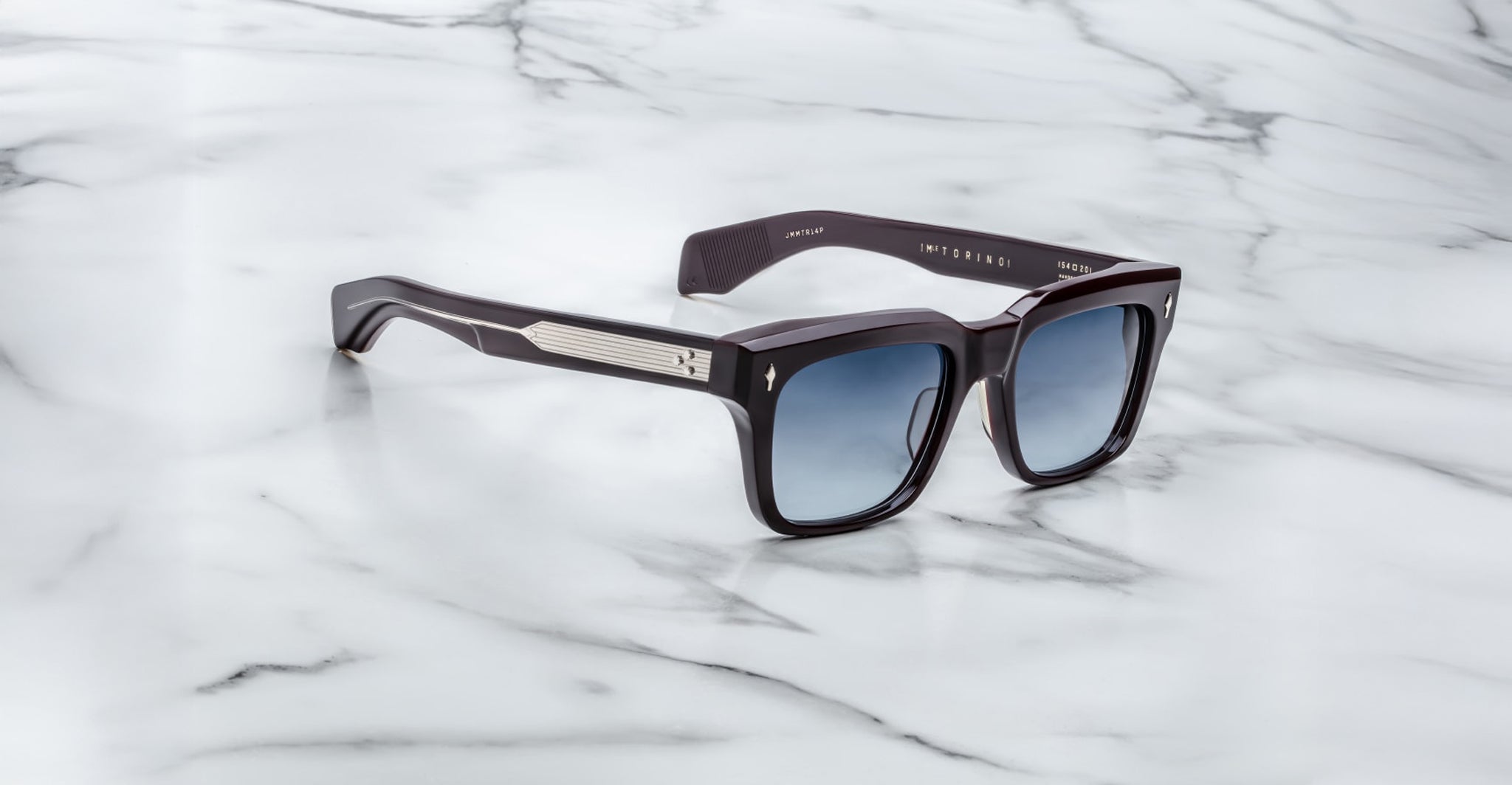 The Jacques Marie Mage Torino by Jacques Marie Mage features black handcrafted acetate rectangular frames, blue gradient lenses, and metallic arm accents, shown on a white marble surface.