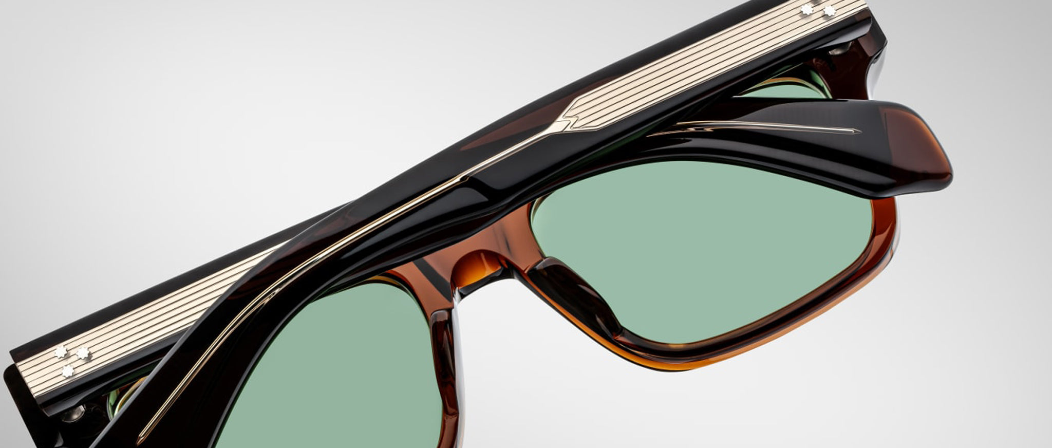 A close-up of Jacques Marie Mage Torino handcrafted acetate eyewear featuring green tinted lenses and dark, glossy frames with metallic and striped temple detailing inspired by bold rectangular designs.
