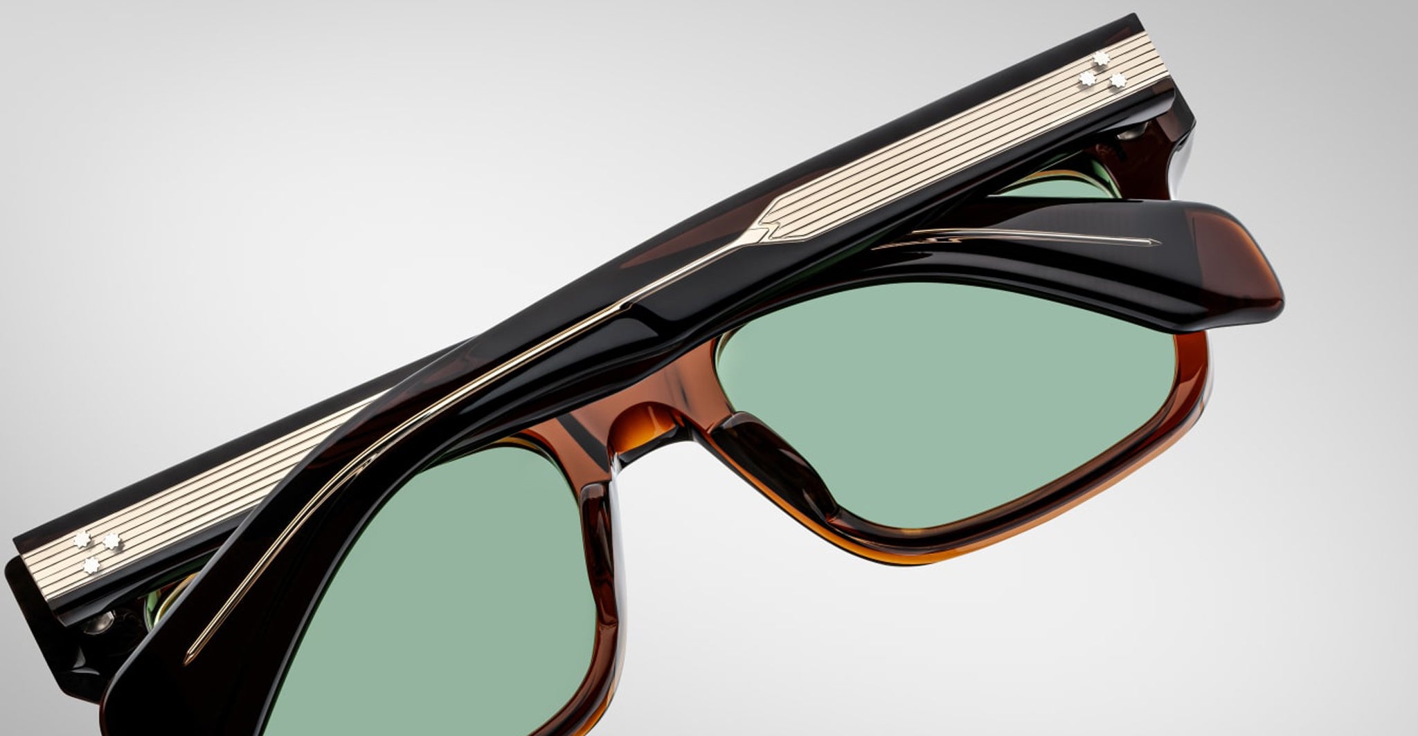 A close-up of Jacques Marie Mage Torino handcrafted acetate eyewear featuring green tinted lenses and dark, glossy frames with metallic and striped temple detailing inspired by bold rectangular designs.