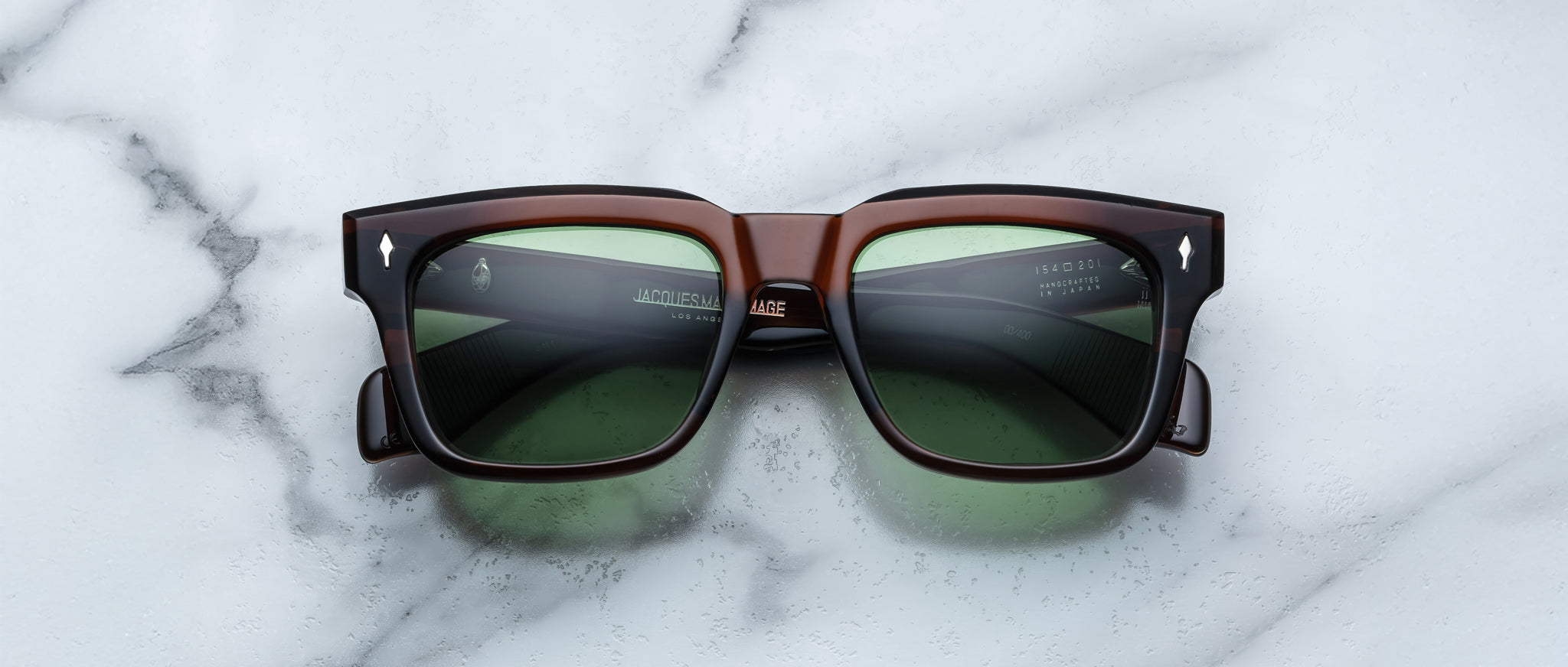 The Jacques Marie Mage Torino, bold rectangular eyeglass frames in dark brown handcrafted acetate with green lenses, are displayed on a white marble surface.