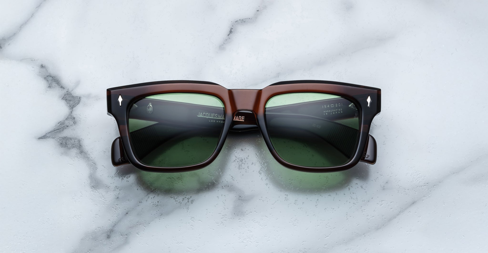 The Jacques Marie Mage Torino, bold rectangular eyeglass frames in dark brown handcrafted acetate with green lenses, are displayed on a white marble surface.