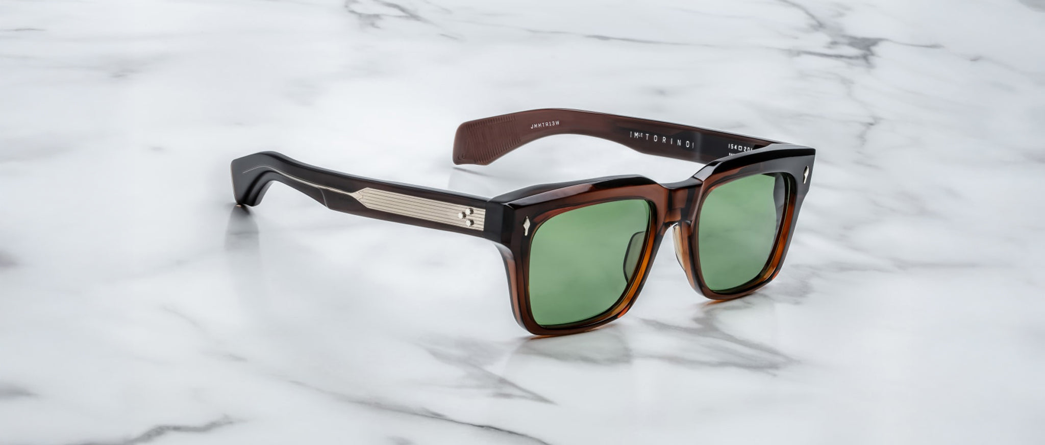 These bold square brown Jacques Marie Mage Torino sunglasses feature green lenses and metallic temple accents, showcasing handcrafted acetate frames displayed elegantly on a white marble surface.