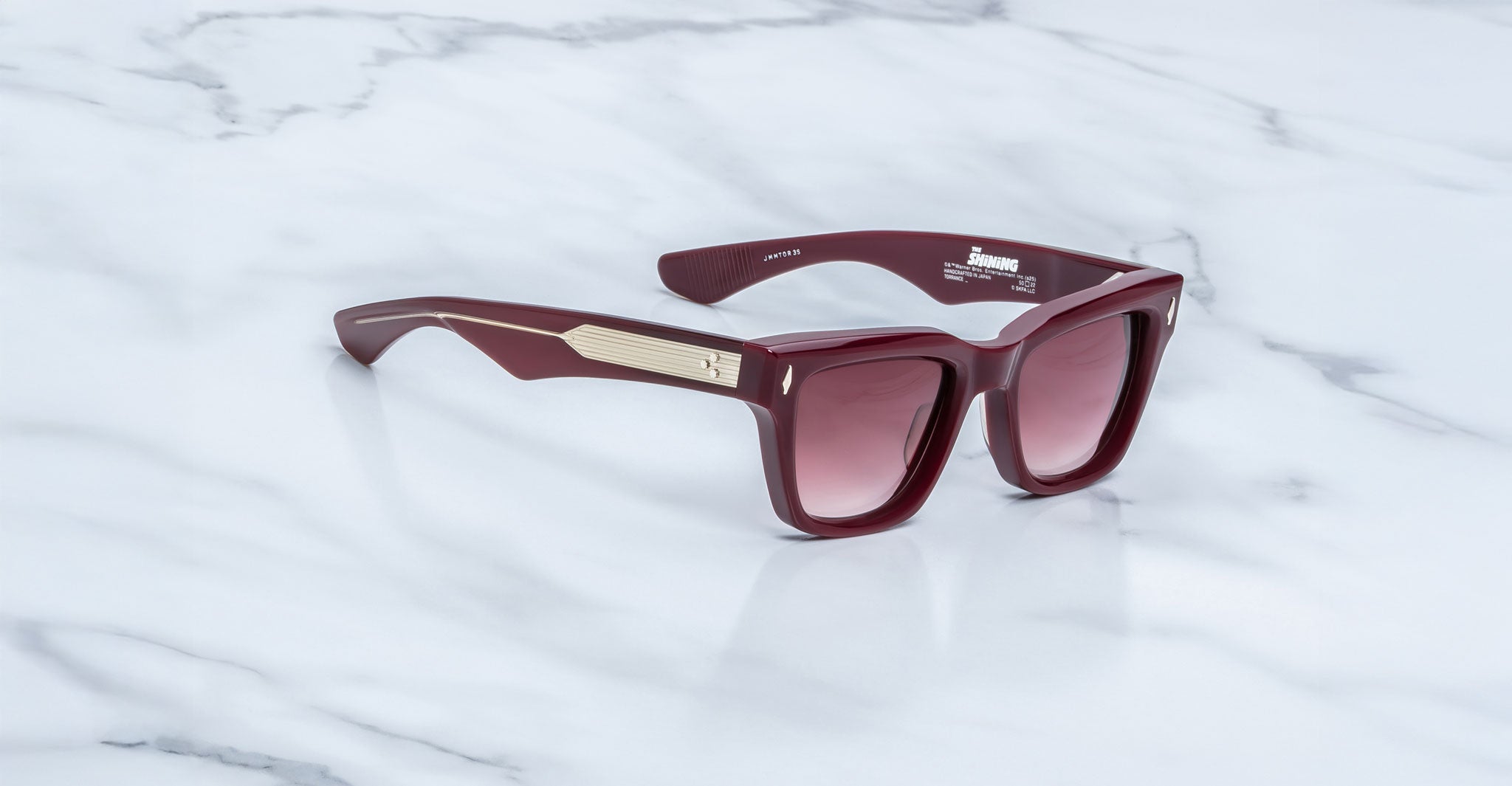 A pair of glossy maroon Jacques Marie Mage Torrance sunglasses with gradient lenses and gold temple detailing, from the limited-edition rectangular The Shining Collection, displayed on a white marble surface.