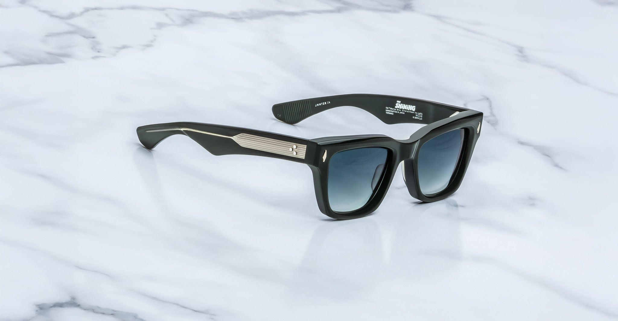 A pair of Jacques Marie Mage Torrance black square sunglasses, from The Shining Collection, featuring limited-edition rectangular frames with dark gradient lenses and metallic temple detailing, resting on a white marble surface.