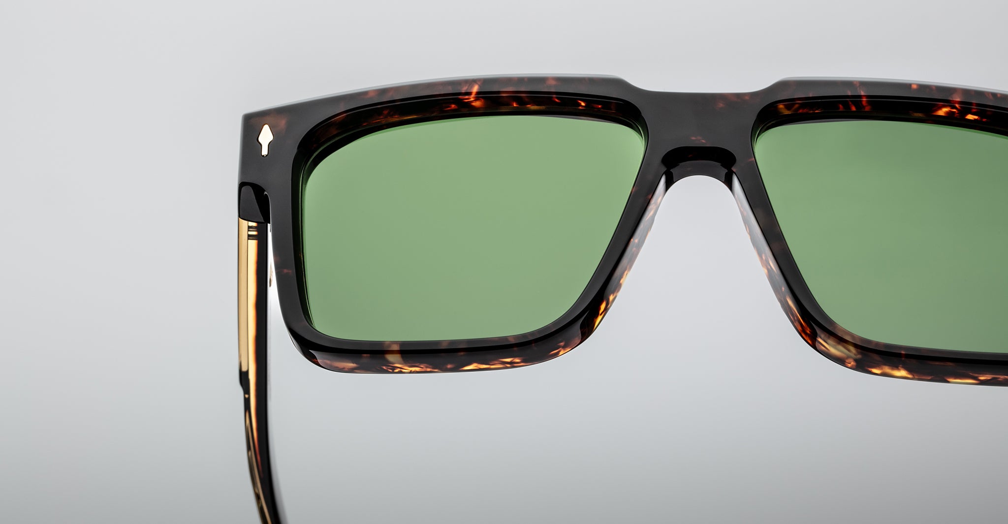 Close-up of a pair of sunglasses with a tortoiseshell frame, green tinted lenses, and gold detailing on the temple, set against a light gray background.