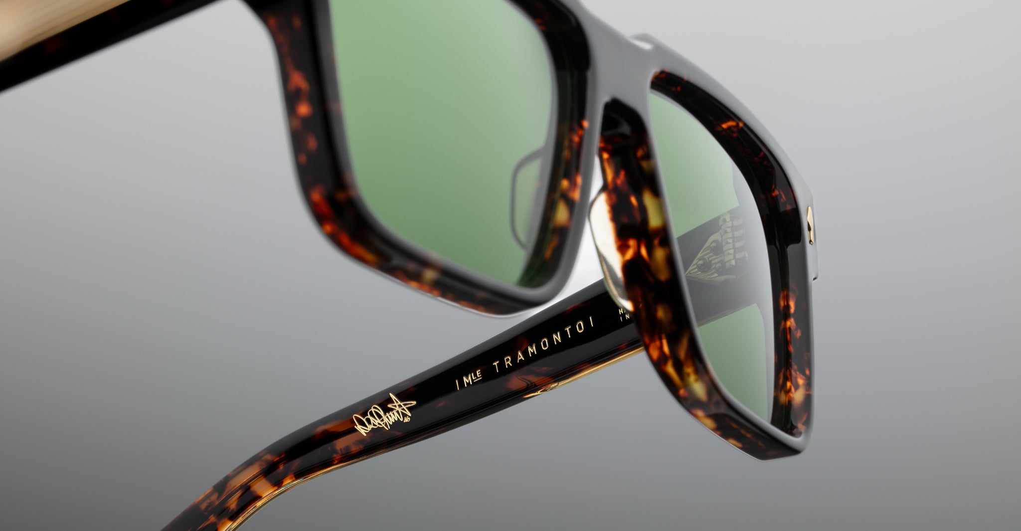 Close-up of tortoiseshell sunglasses with green lenses, showing detailed frame texture and the inscription IME TRAMONTO I in gold on the arm. The background is a smooth, neutral gray.