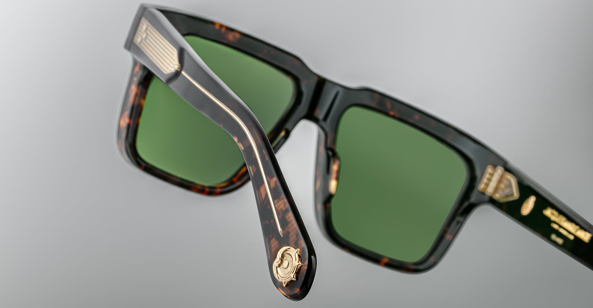 Close-up of stylish tortoiseshell sunglasses with green lenses, gold detailing on the arms, and a gold emblem on the hinge, set against a plain, light gray background.