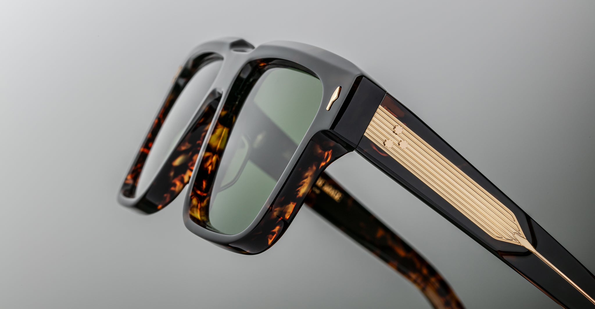 Close-up of stylish sunglasses with thick tortoiseshell frames, green tinted lenses, and gold decorative accents on the temples, set against a minimal gray background.