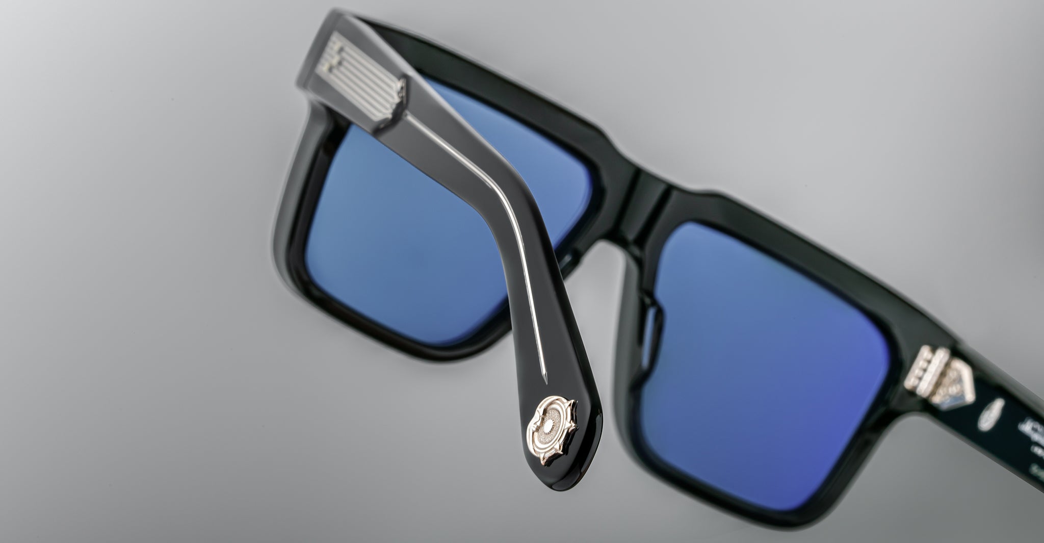 Close-up of black rectangular sunglasses with blue-tinted lenses, featuring silver decorative accents on the frame and temple, set against a plain gray background.