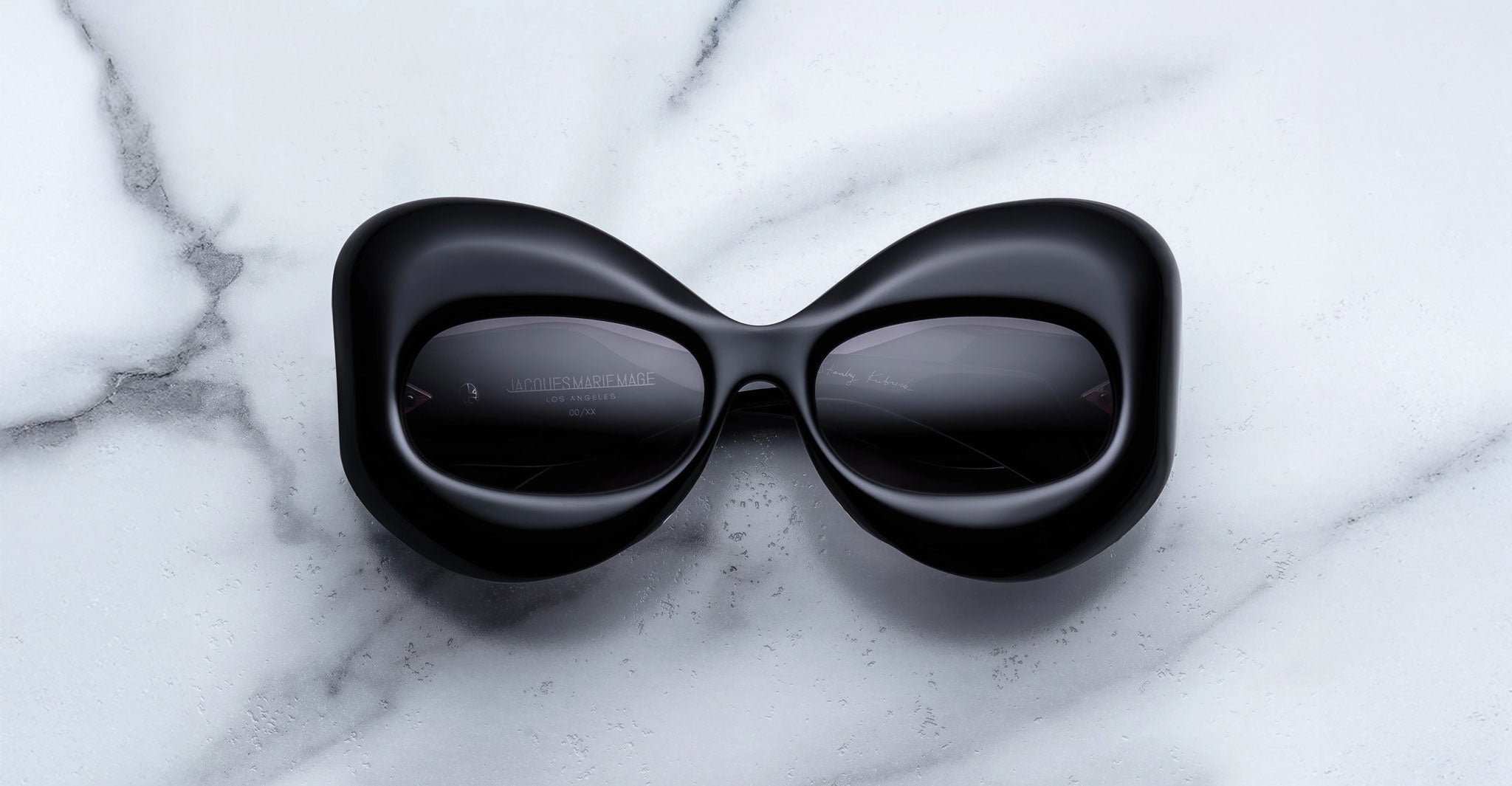 A pair of oversized, black Jacques Marie Mage Twins sunglasses from The Shining Collection, inspired by Stanley Kubrick, rests on white marble with gray veining—distinctive limited-edition eyewear for the discerning.