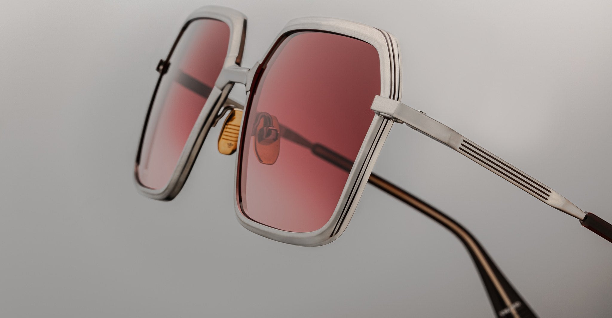 Close-up of Jacques Marie Mage Ugo sunglasses featuring large square frames, metallic silver rims, and pink-tinted lenses. Visible nose pads and temple arms are set against a plain, light background.