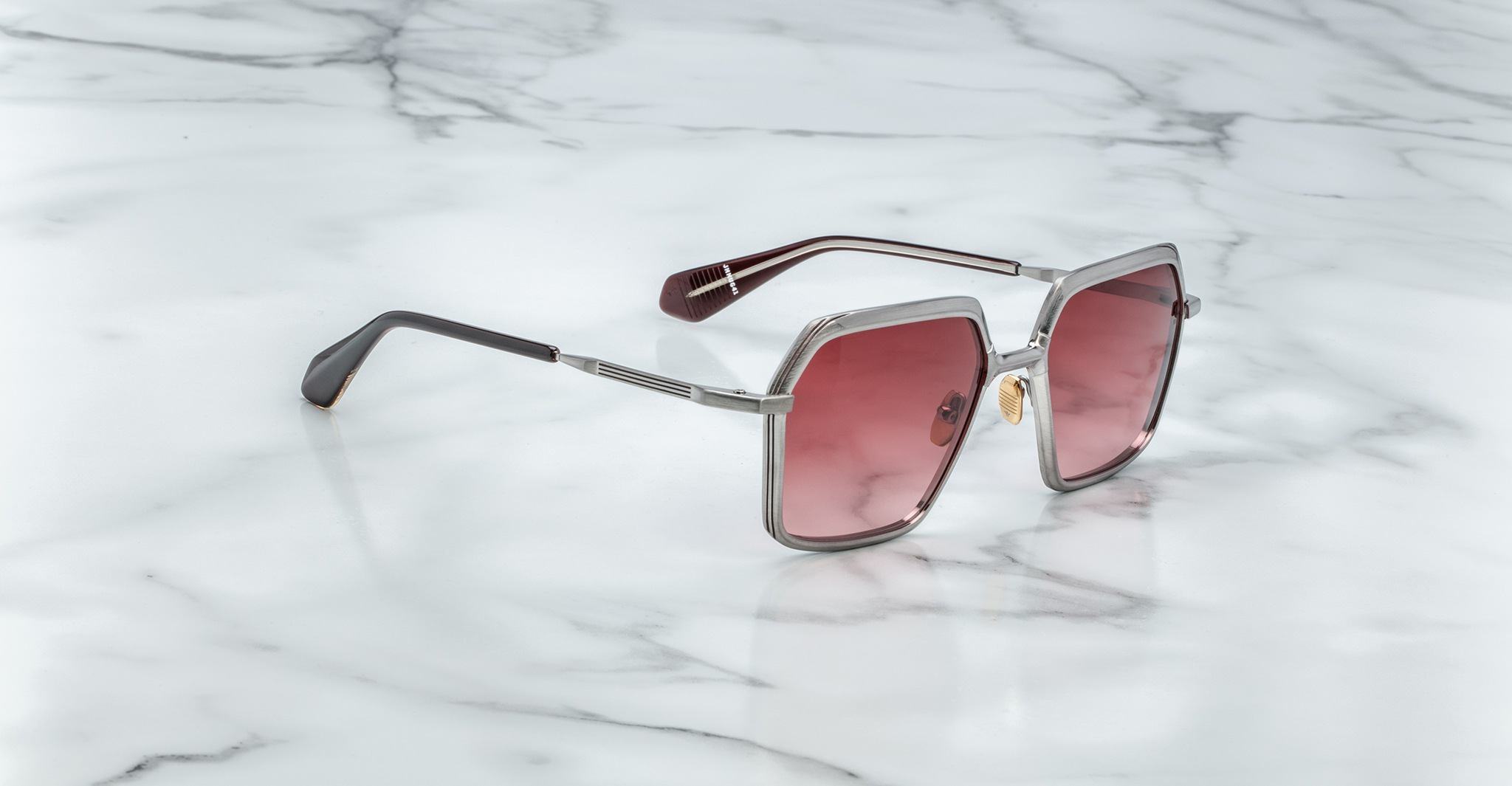 The Jacques Marie Mage Ugo sunglasses, featuring square frames, pink-tinted lenses, and silver rims, rest on a white marble surface.