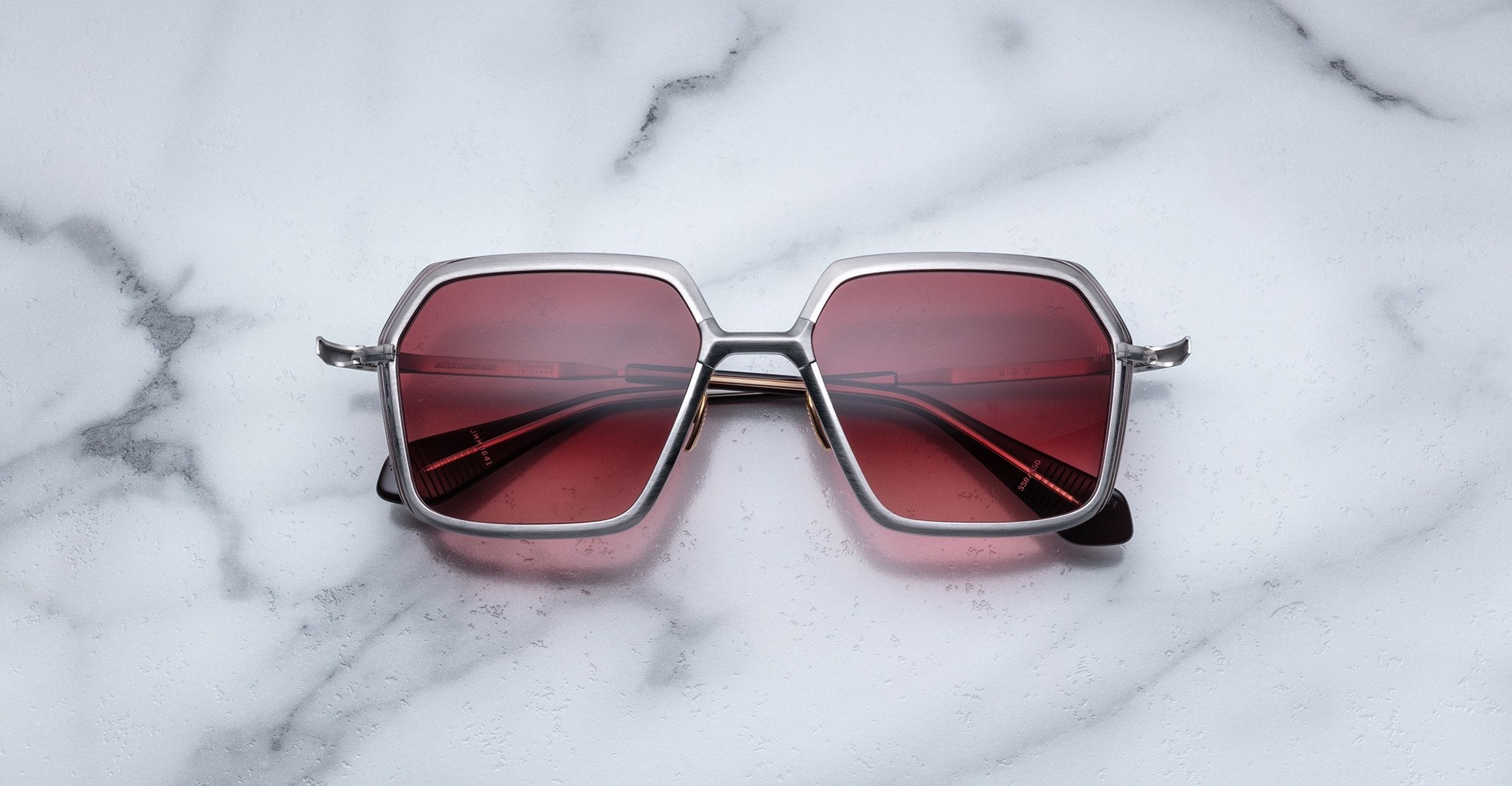 The Jacques Marie Mage Ugo sunglasses, featuring square silver frames and red-tinted lenses, are displayed on a white marble surface.