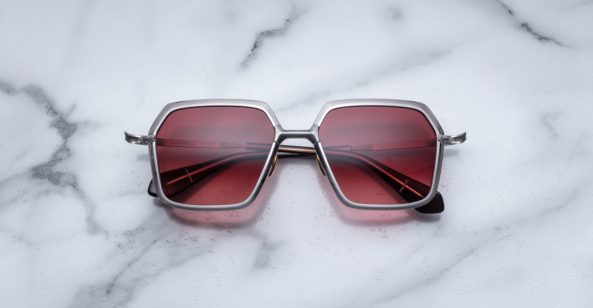 The Jacques Marie Mage Ugo sunglasses, featuring square silver frames and red-tinted lenses, are displayed on a white marble surface.