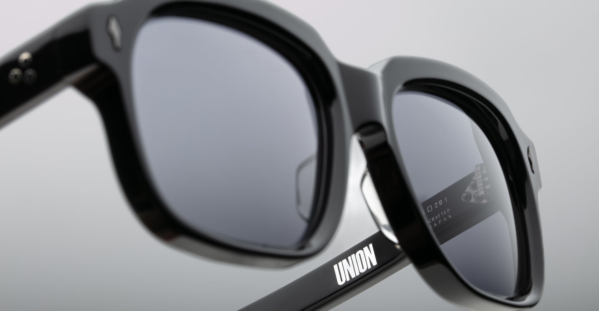 Close-up of Jacques Marie Mage Union black sunglasses with thick frames. UNION is printed in white on the temple arm, and numbers and text reflect in the lens. The background is neutral and out of focus.