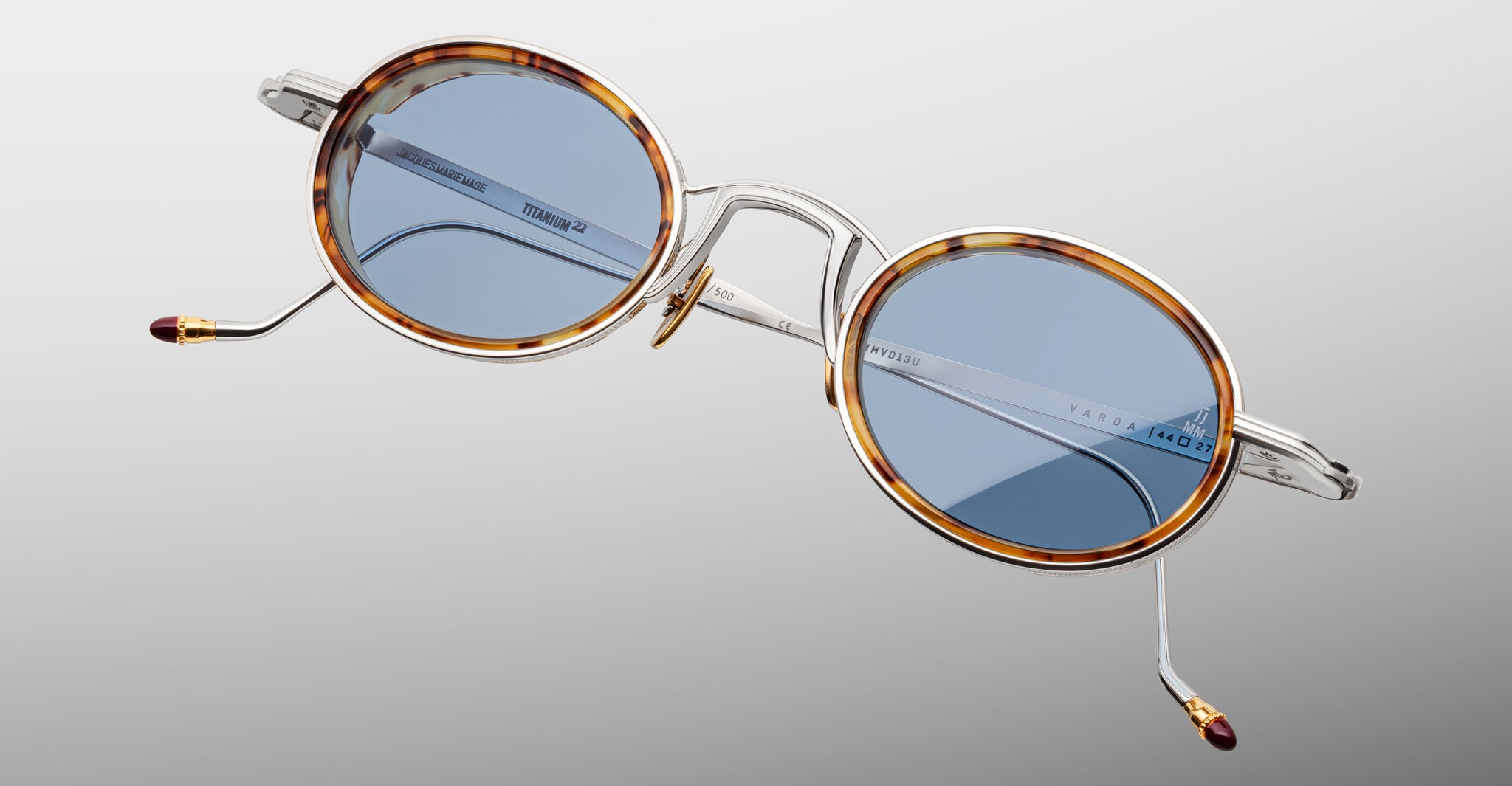 The Jacques Marie Mage Varda features round eyeglasses with tortoiseshell-patterned rims, silver metal arms, blue-tinted lenses, and gold and burgundy accents on the arm tips. Set against a plain gradient grey background.