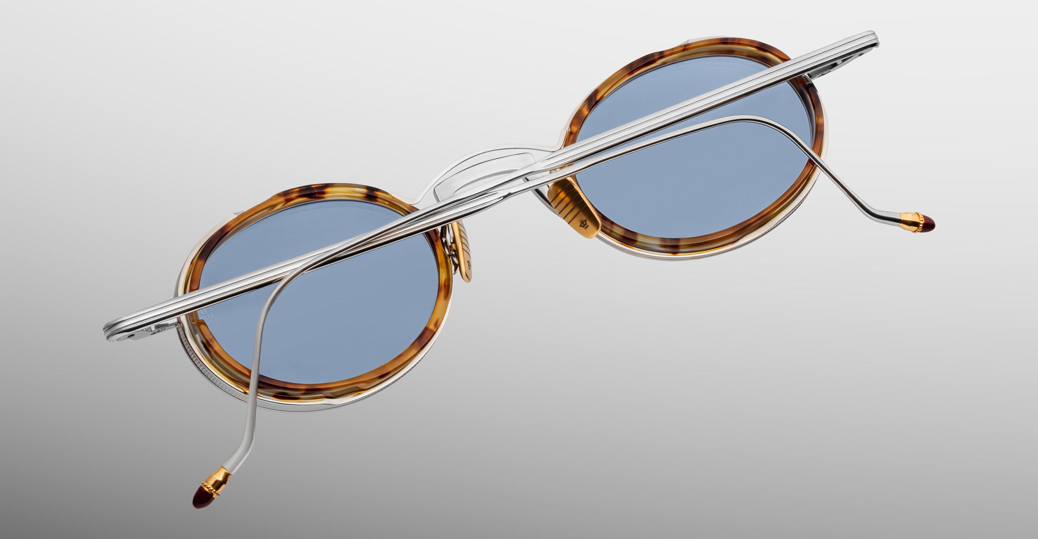 The Jacques Marie Mage Varda round sunglasses feature blue lenses, tortoiseshell frames, and thin silver metal arms, displayed from the back against a light gray gradient background.
