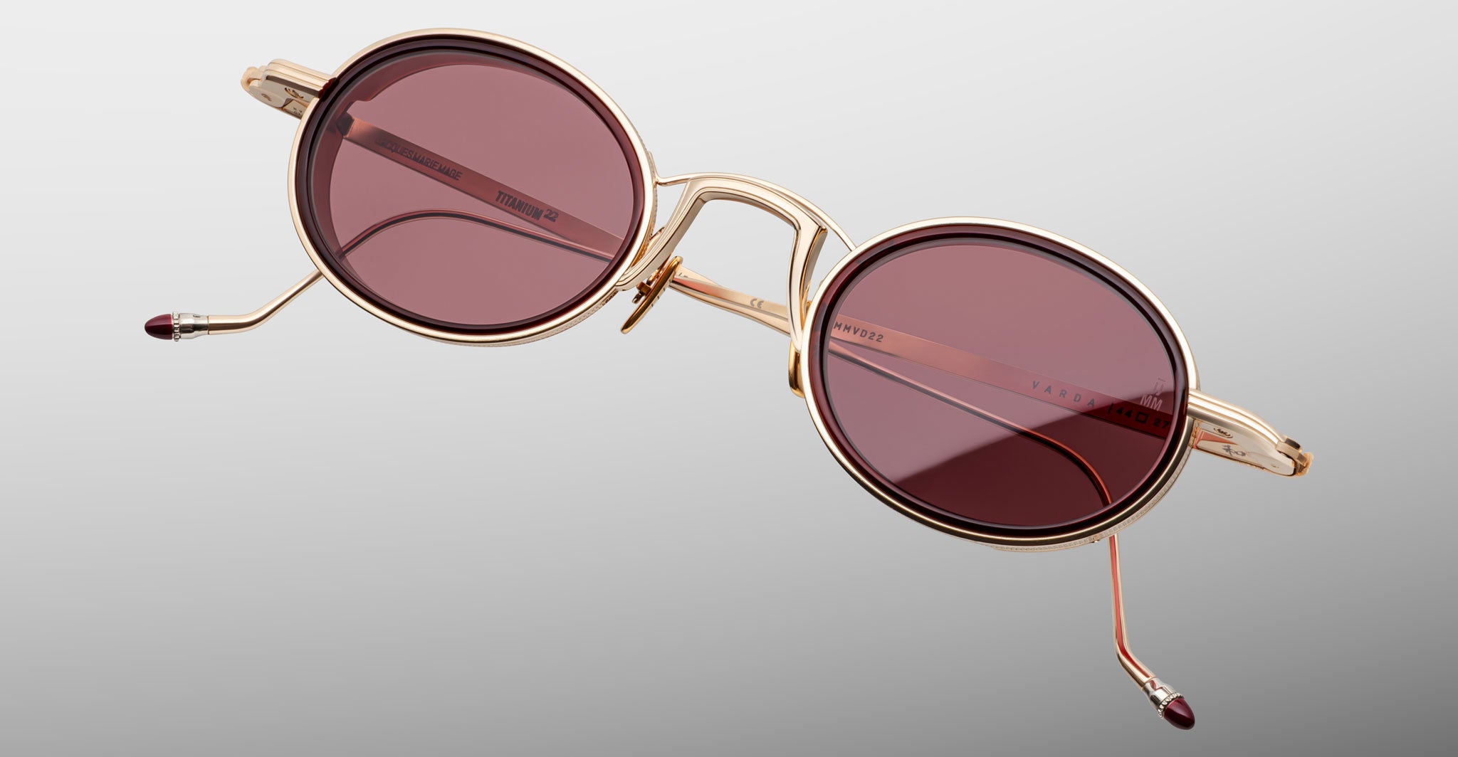 The Jacques Marie Mage Varda sunglasses are featured with round gold metal frames, dark red-tinted lenses, and matching gold temples with red tips, displayed against a plain gray background.