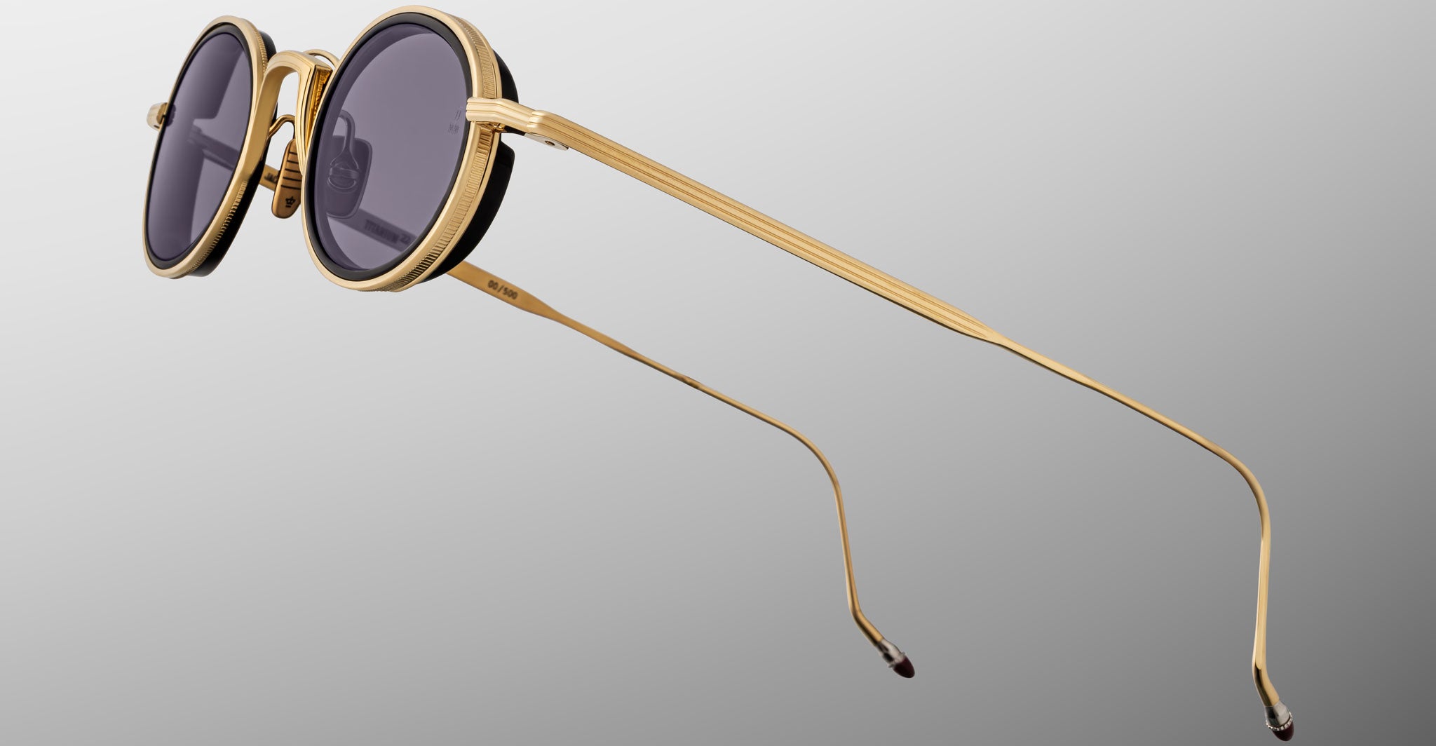 The Jacques Marie Mage Varda sunglasses feature round gold metal frames with black rims and dark tinted lenses, displayed at an angle on a neutral gray background. The thin temples are slightly curved at the tips.