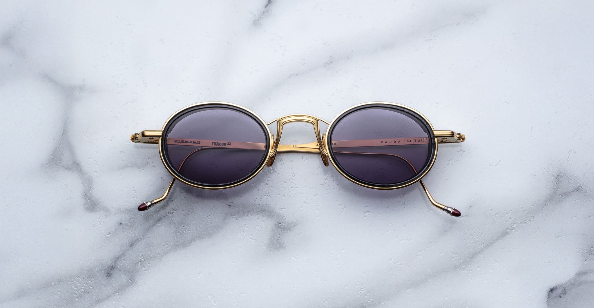 The Jacques Marie Mage Varda sunglasses, featuring round gold frames and dark lenses with subtle text on the lens and inner arm, are displayed on a white marble surface with visible arms.