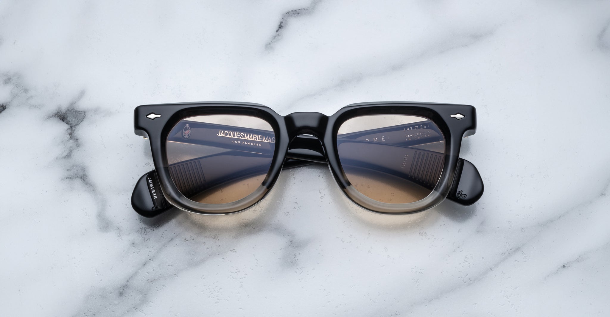A pair of Jacques Marie Mage Vendome sunglasses by Jacques Marie Mage, featuring black rims and brown gradient lenses, handcrafted in Japan, is displayed on a white marble surface.