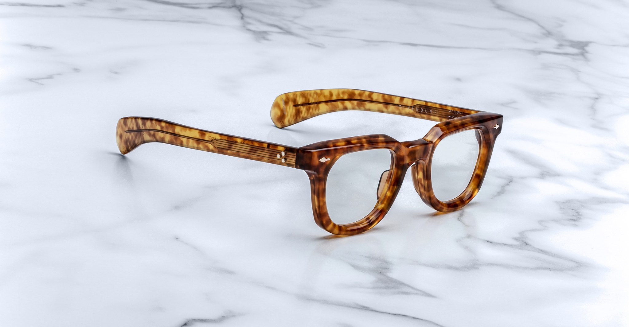 Jacques Marie Mage Vendome optical frames, featuring a tortoiseshell pattern and handcrafted in Japan, are displayed on a white marble surface.