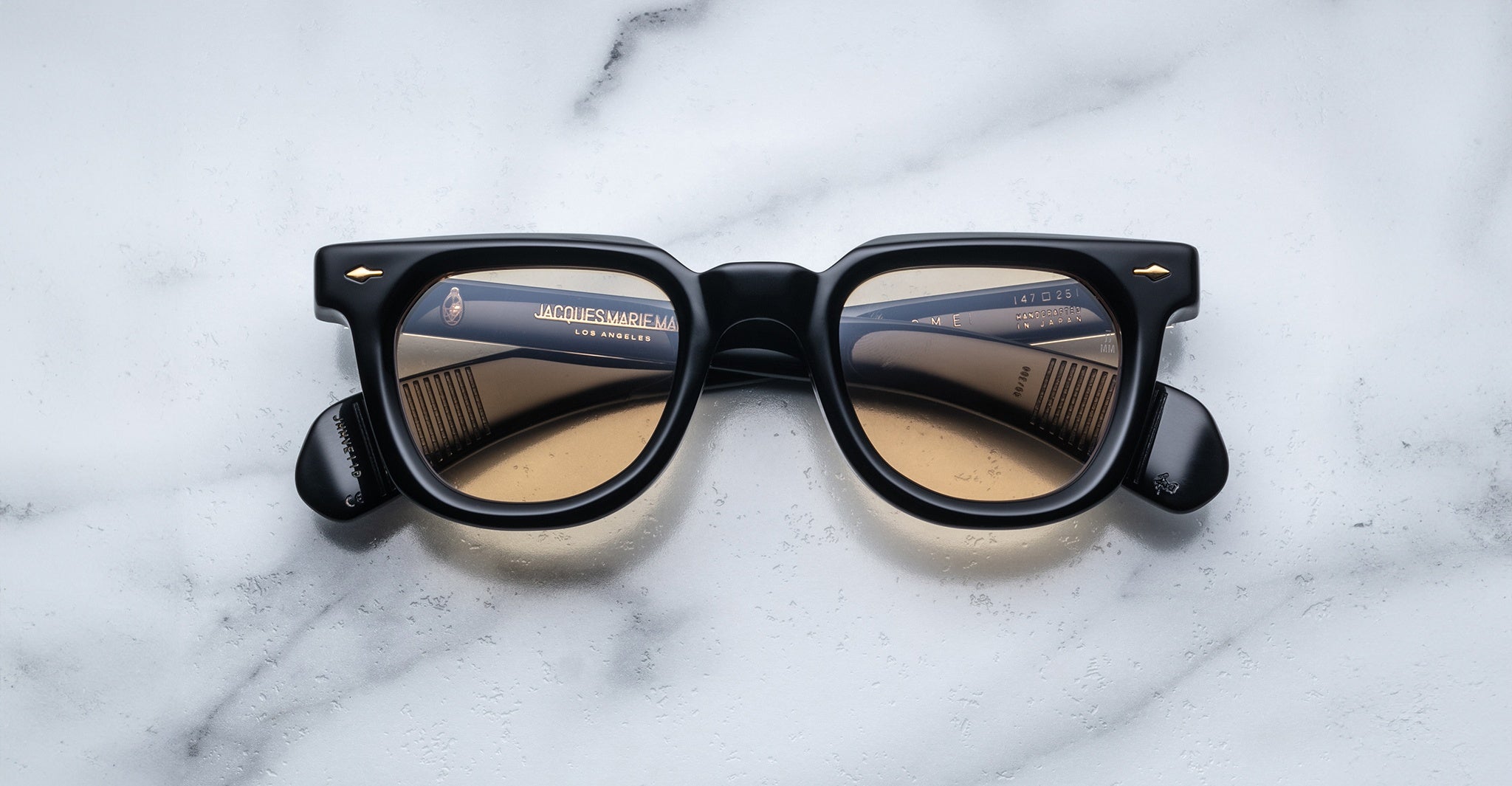 A pair of Jacques Marie Mage Vendome sunglasses with black rims and brown-tinted lenses sits on white marble, the brand name visible inside the arms. Handcrafted in Japan for impeccable quality.