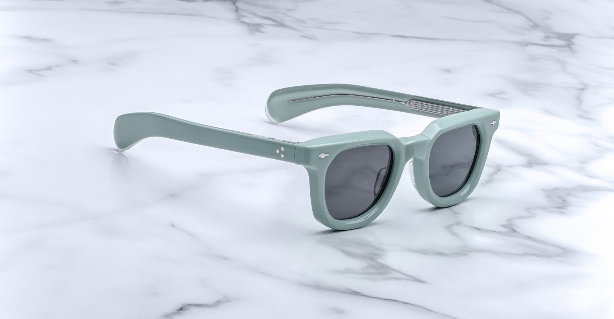 Jacques Marie Mage Vendome sunglasses in pale green with dark lenses rest on white marble, showcasing the brand’s expert Japanese handcrafted craftsmanship.