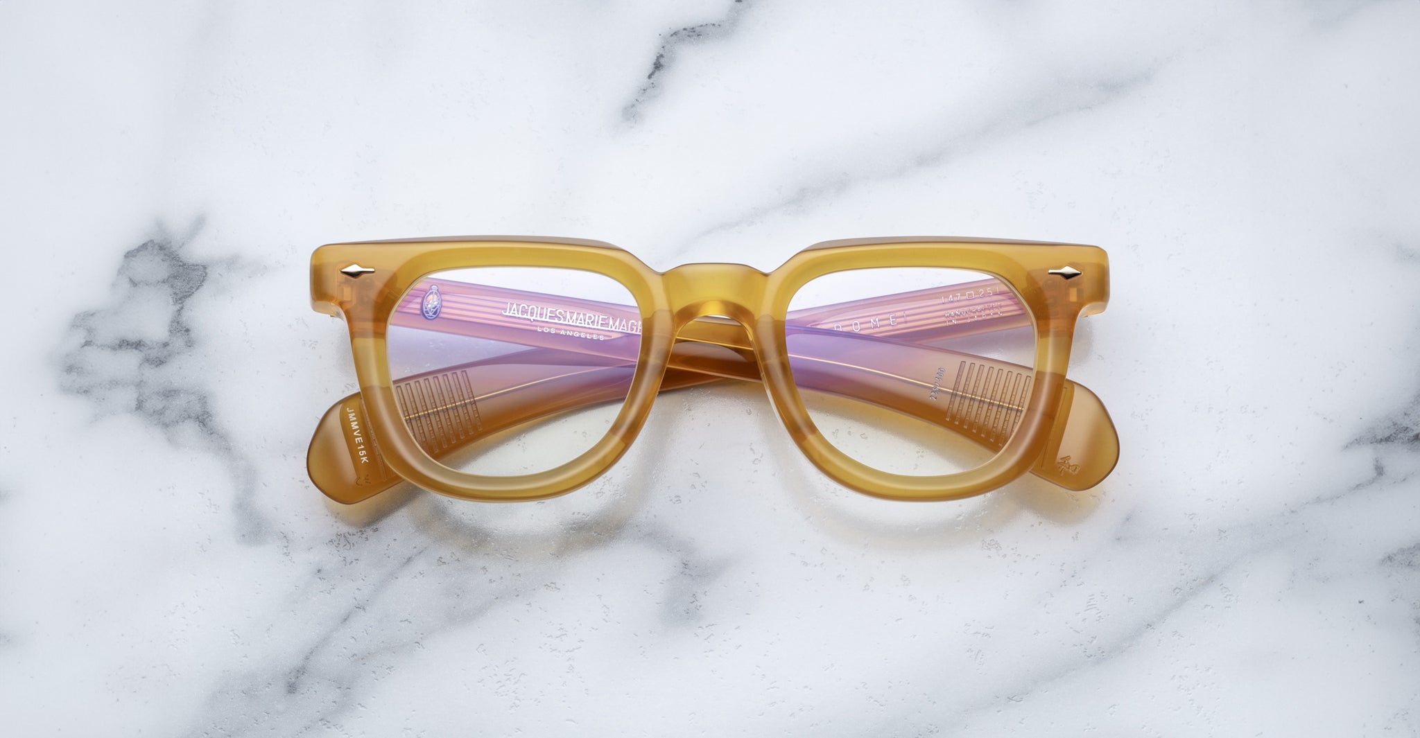 A pair of Jacques Marie Mage Vendome amber optical frames with reflective pink lenses, handcrafted in Japan, rests on a white marble surface.