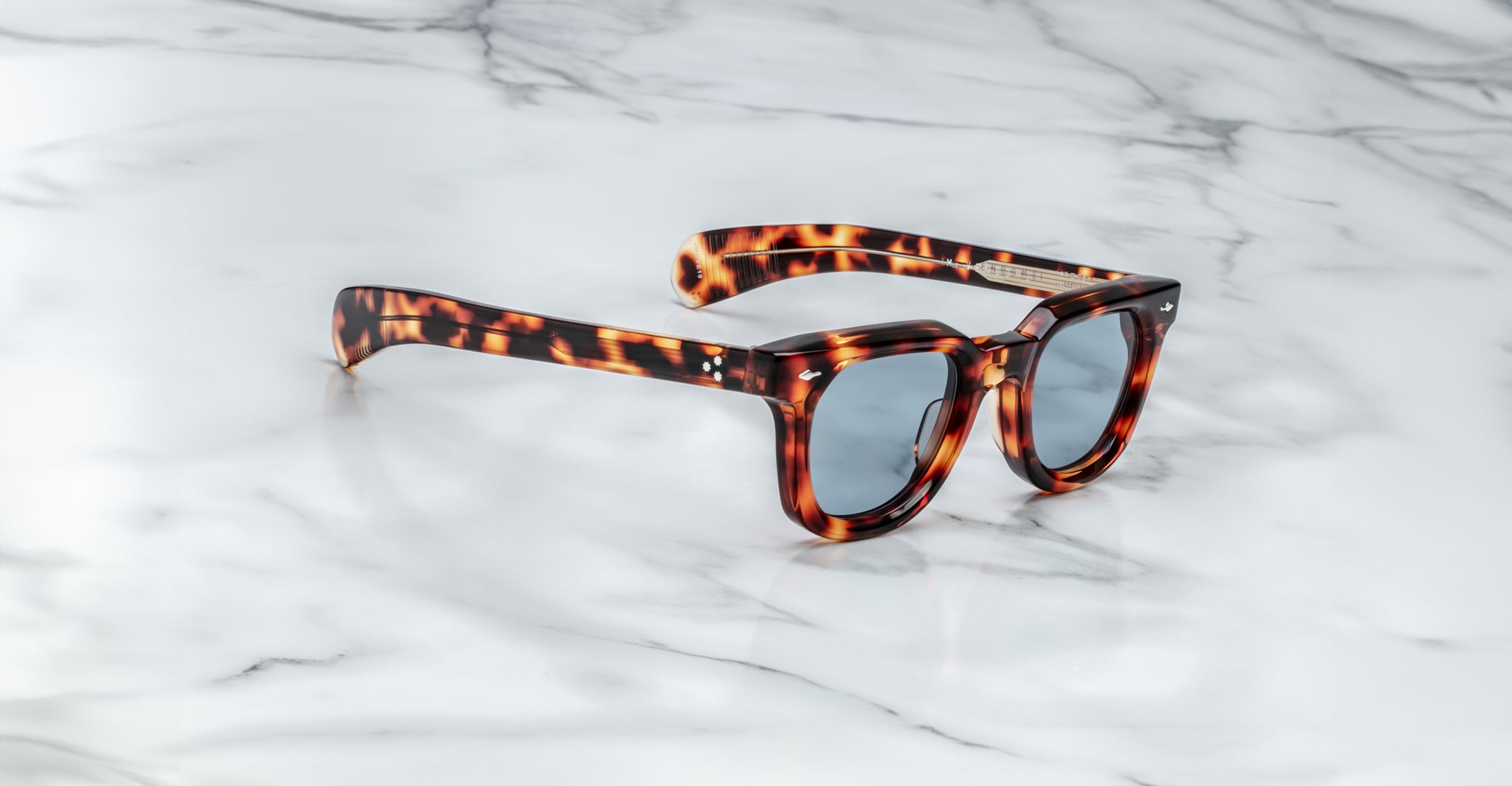 Jacques Marie Mage Vendome sunglasses with a tortoiseshell pattern and blue-tinted lenses rest on white marble, highlighting the handcrafted Japanese optical frames by Jacques Marie Mage.