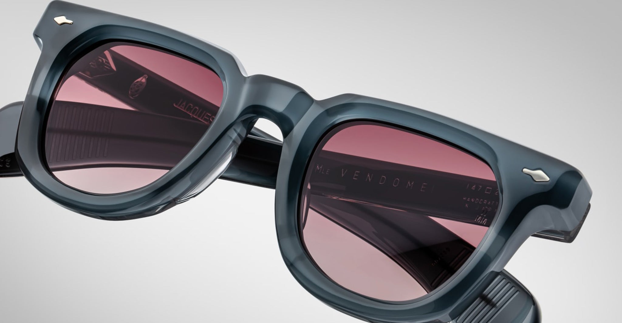 Close-up of Jacques Marie Mage Vendome sunglasses by Jacques Marie Mage, featuring thick frames and pink lenses, handcrafted in Japan, set against a gray background.
