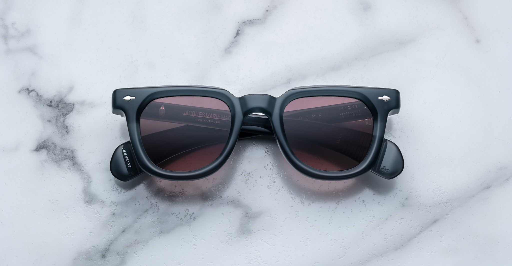 A pair of Jacques Marie Mage Vendome square sunglasses in black with dark lenses, handcrafted in Japan, rests on a white marble surface.