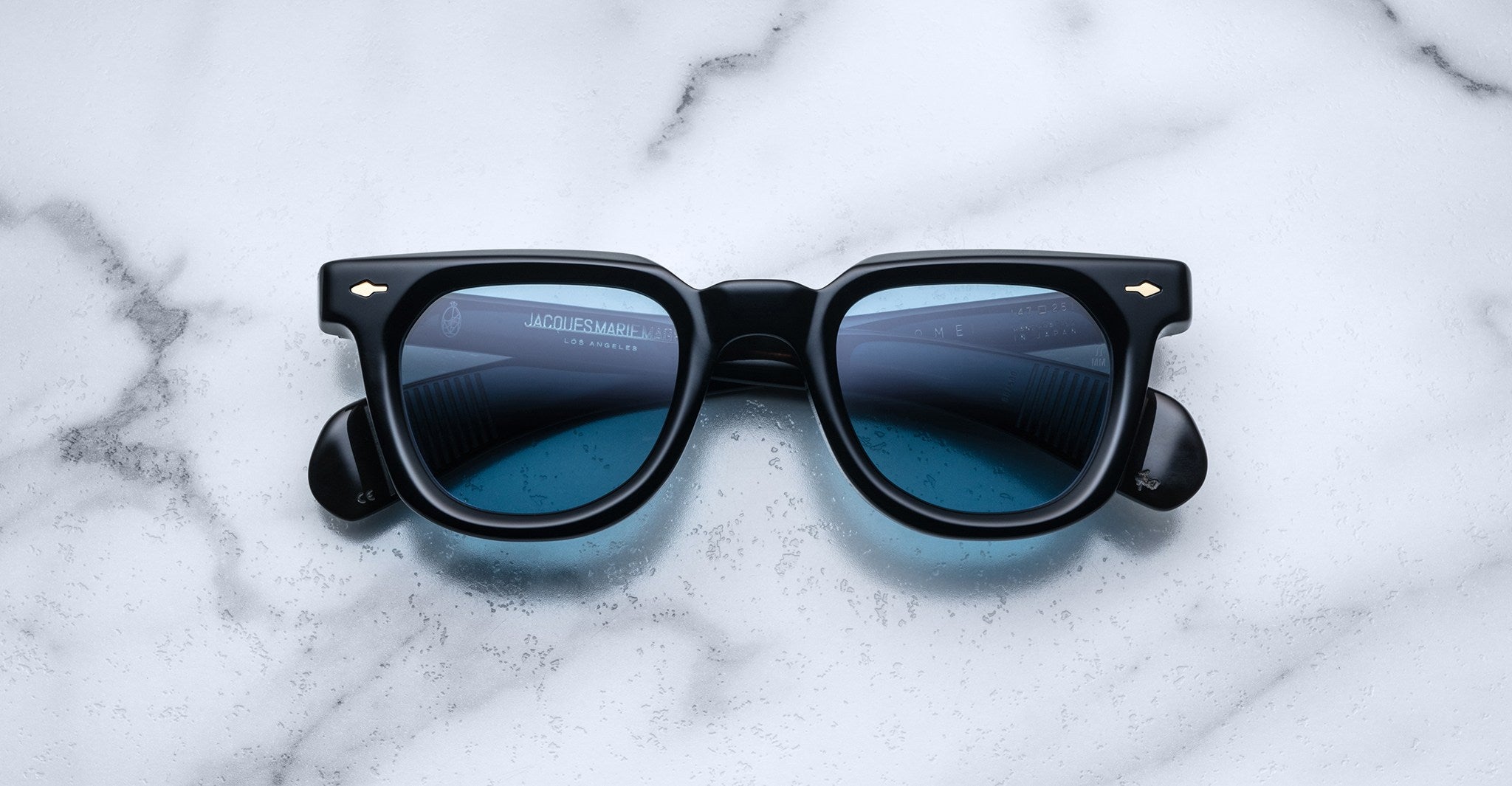 A pair of Jacques Marie Mage Vendome sunglasses by Jacques Marie Mage, featuring black frames and blue tinted lenses, handcrafted in Japan, rests on a white marble surface.
