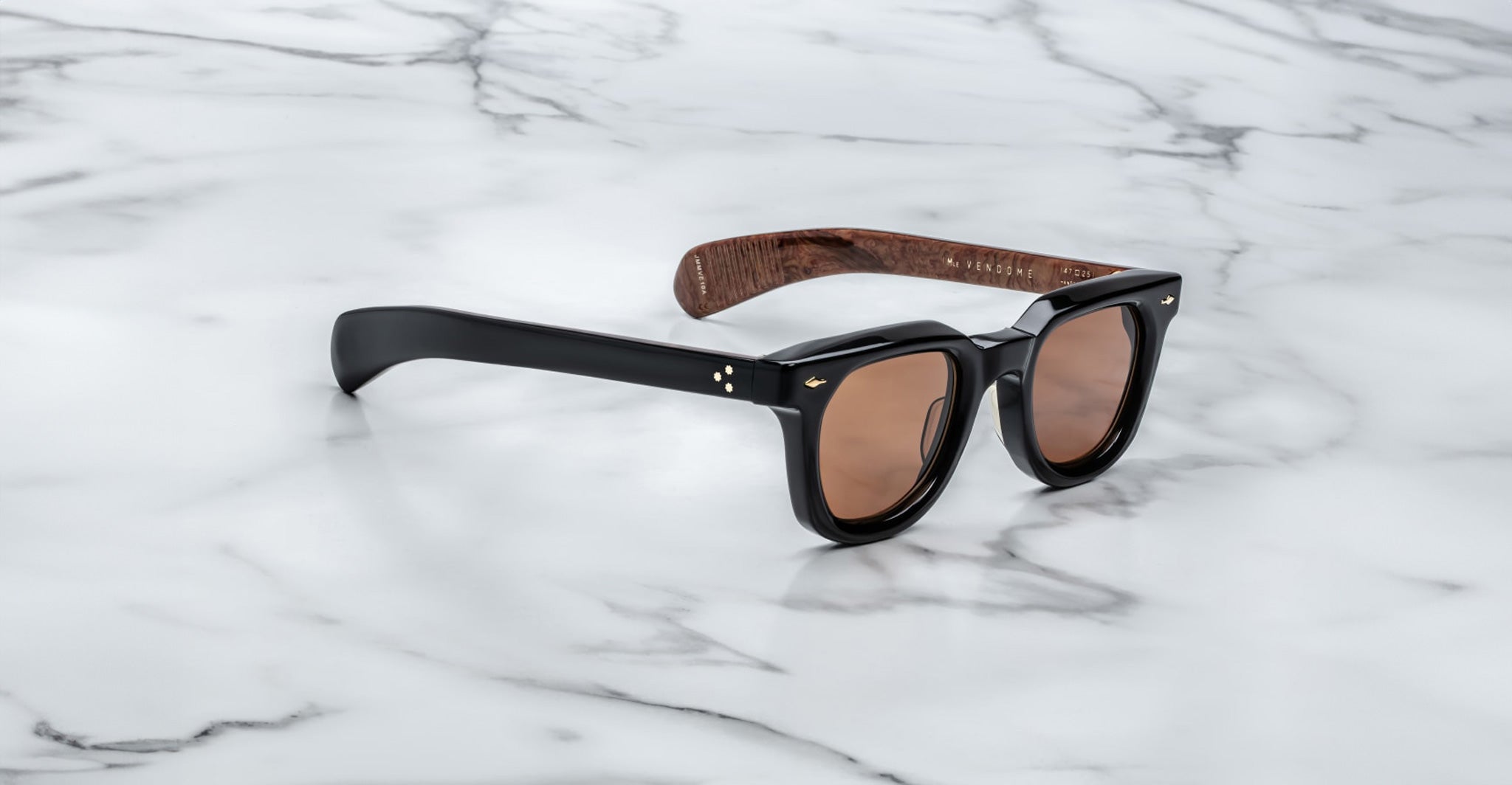 The Jacques Marie Mage Vendome sunglasses by Jacques Marie Mage feature black frames, brown lenses, and wood-textured inner arms. Handcrafted in Japan, they are displayed on a white marble surface.