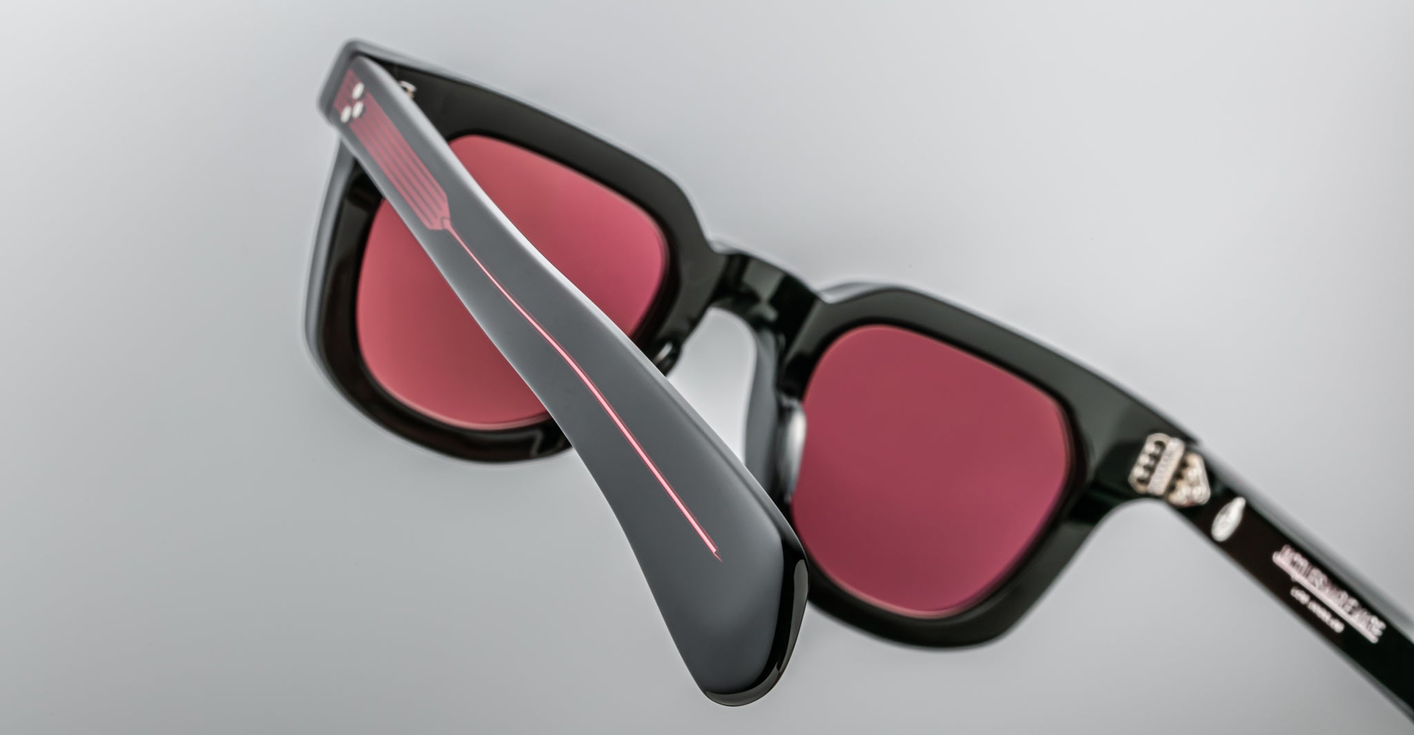 Close-up of Jacques Marie Mage Vendome sunglasses by Jacques Marie Mage, featuring black frames, red-tinted lenses, and a thin red stripe on the arms, meticulously handcrafted in Japan against a plain gray background.